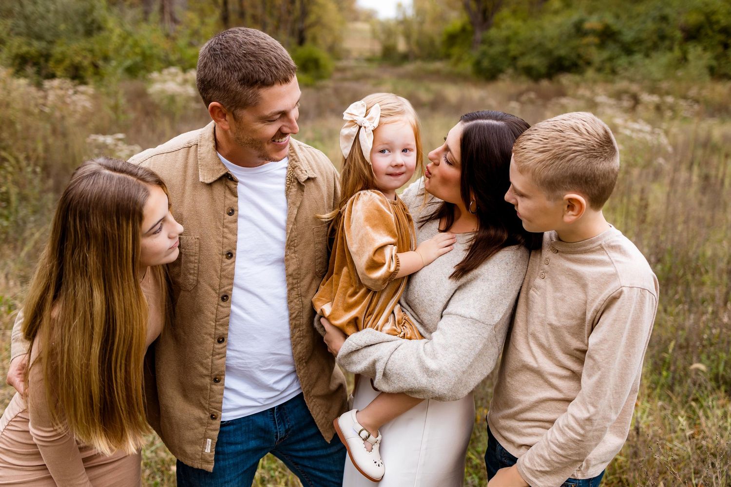 Family wearing coordinated neutral tones embraces while sitting in a golden autumn field at sunset.