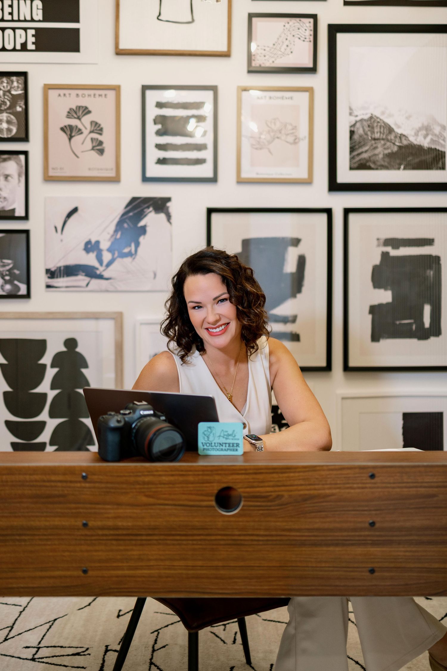 Professional photographer peeks over a wooden desk in a studio space decorated with framed artwork on the wall.