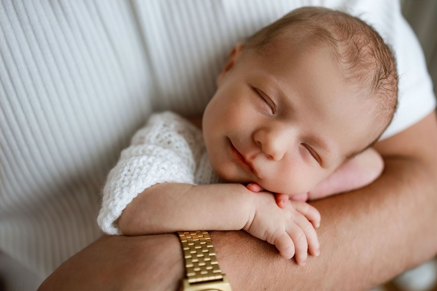 Sleeping newborn baby smiling peacefully while resting on parent's arm wearing a gold watch.