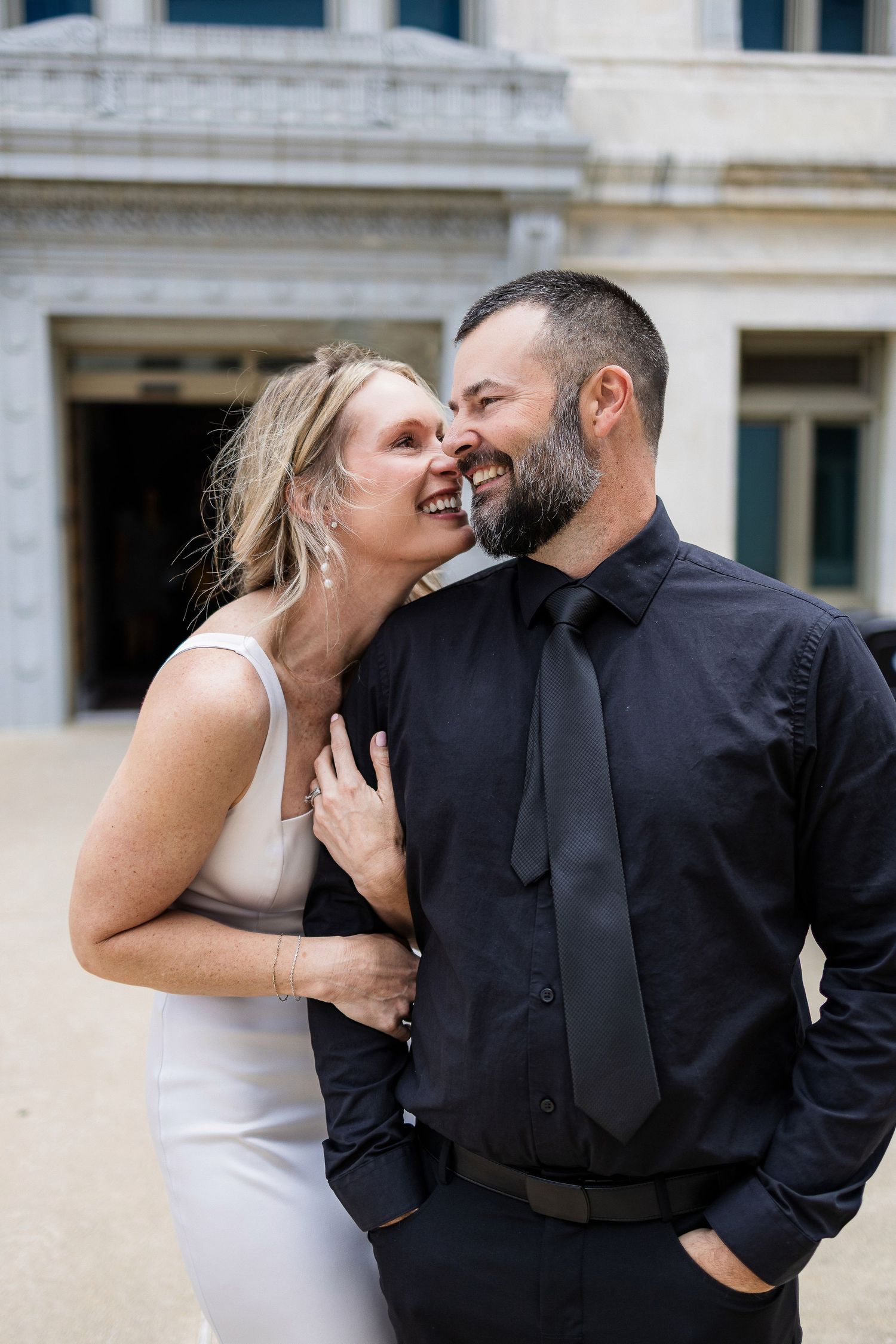 A joyful couple sharing a candid laughing moment outside a building, wearing black and white formal attire.