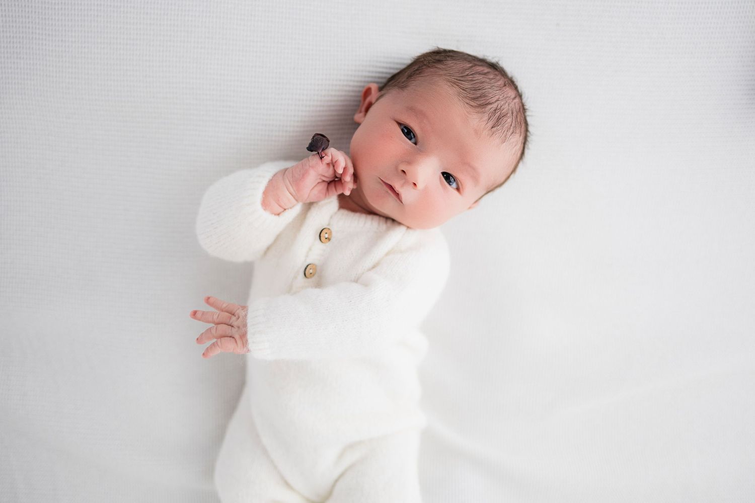 Newborn baby wearing white knit outfit lying on soft white backdrop during professional photo session.
