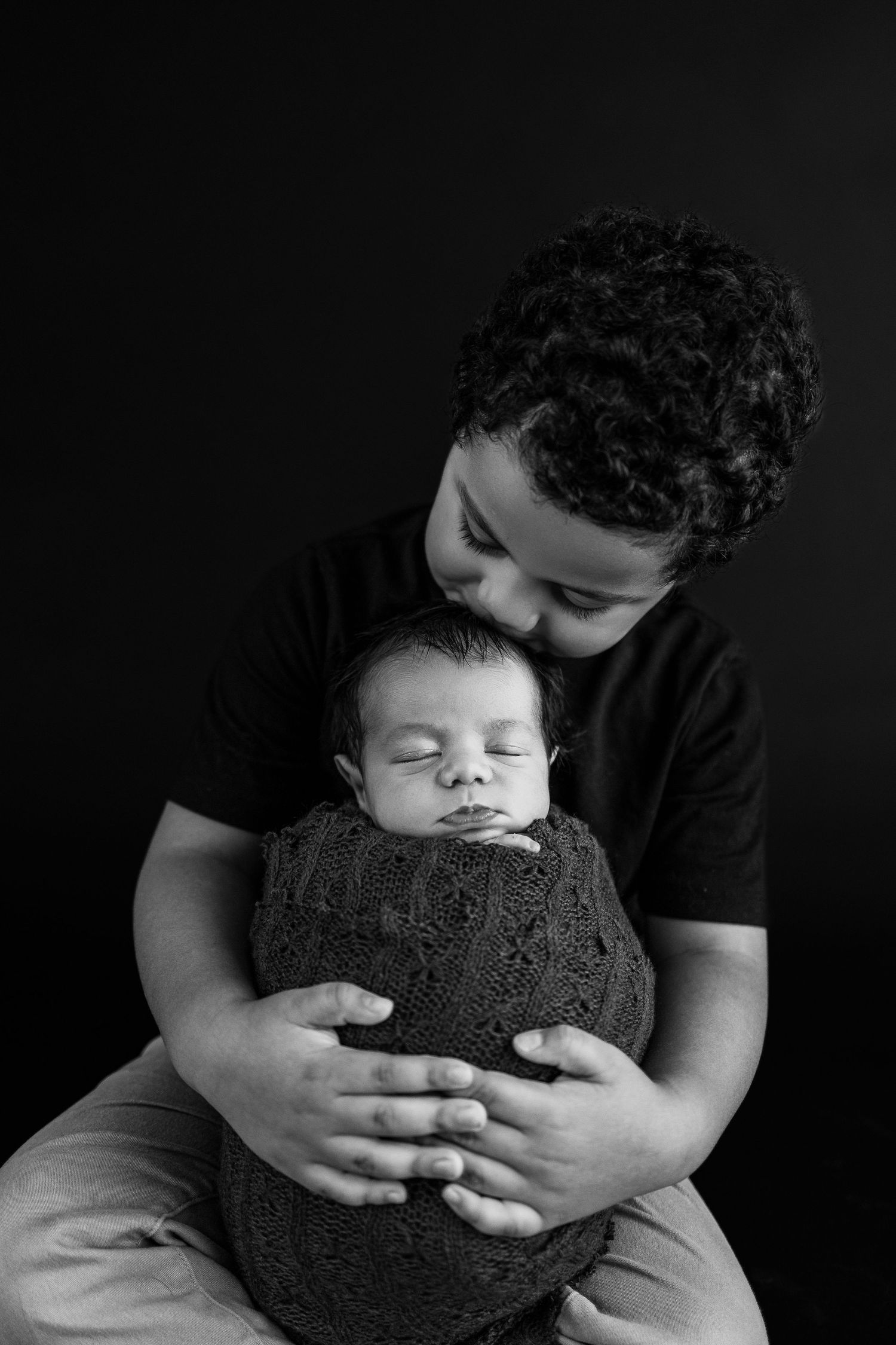 Black and white portrait of two siblings, with older child gently cradling swaddled sleeping newborn against dark background.