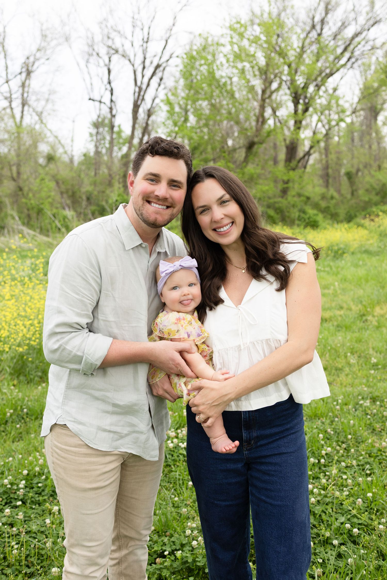 Happy family of three pose together in spring meadow with yellow wildflowers.