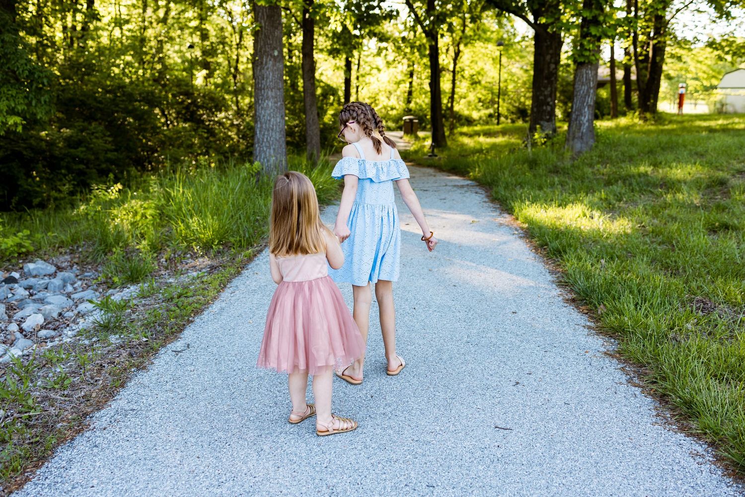 Two children walking hand in hand down a sunlit gravel path surrounded by greenery.