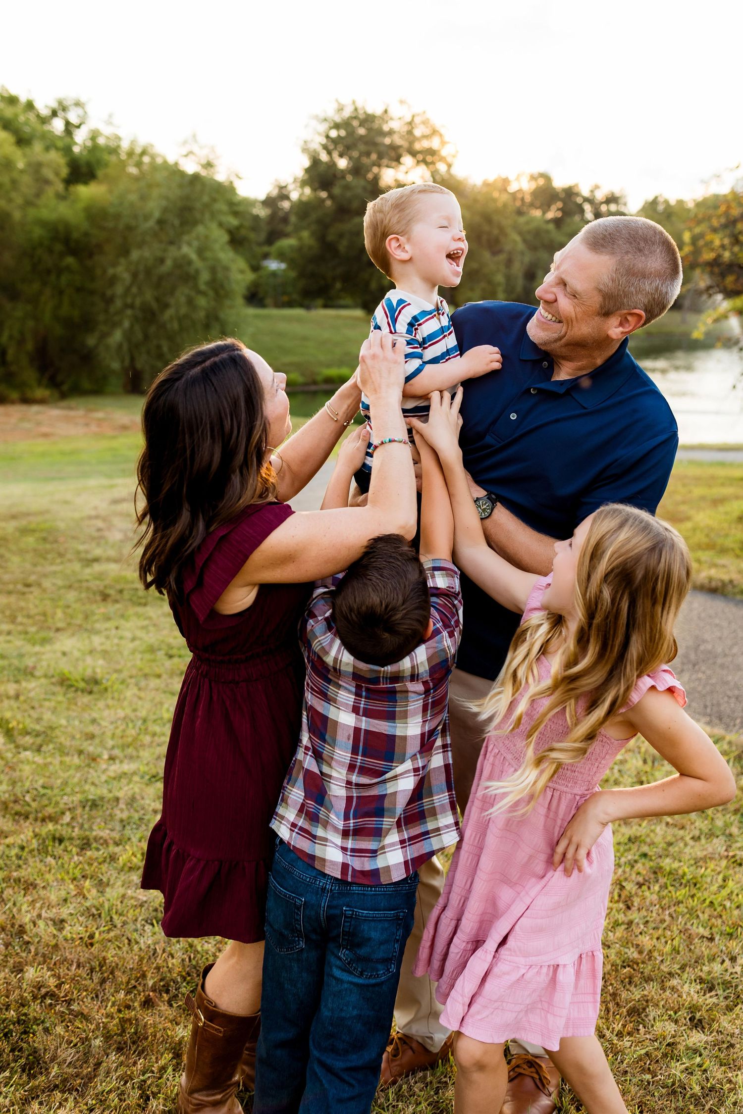 Family of three playing and laughing together outdoors during golden hour sunset in a grassy field by the water.