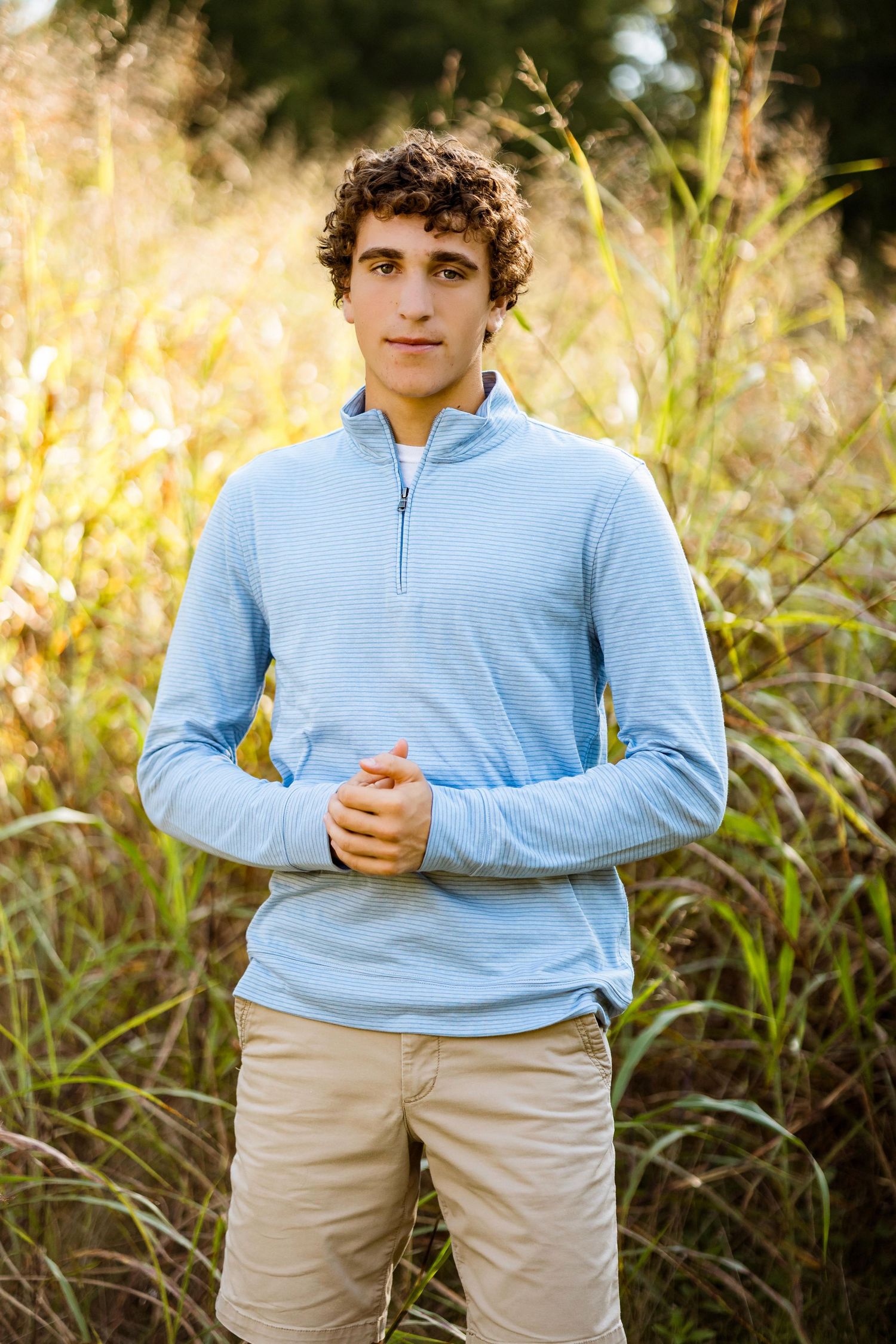 A person in a light blue quarter-zip pullover and khaki pants stands in front of tall grass on a sunny day.
