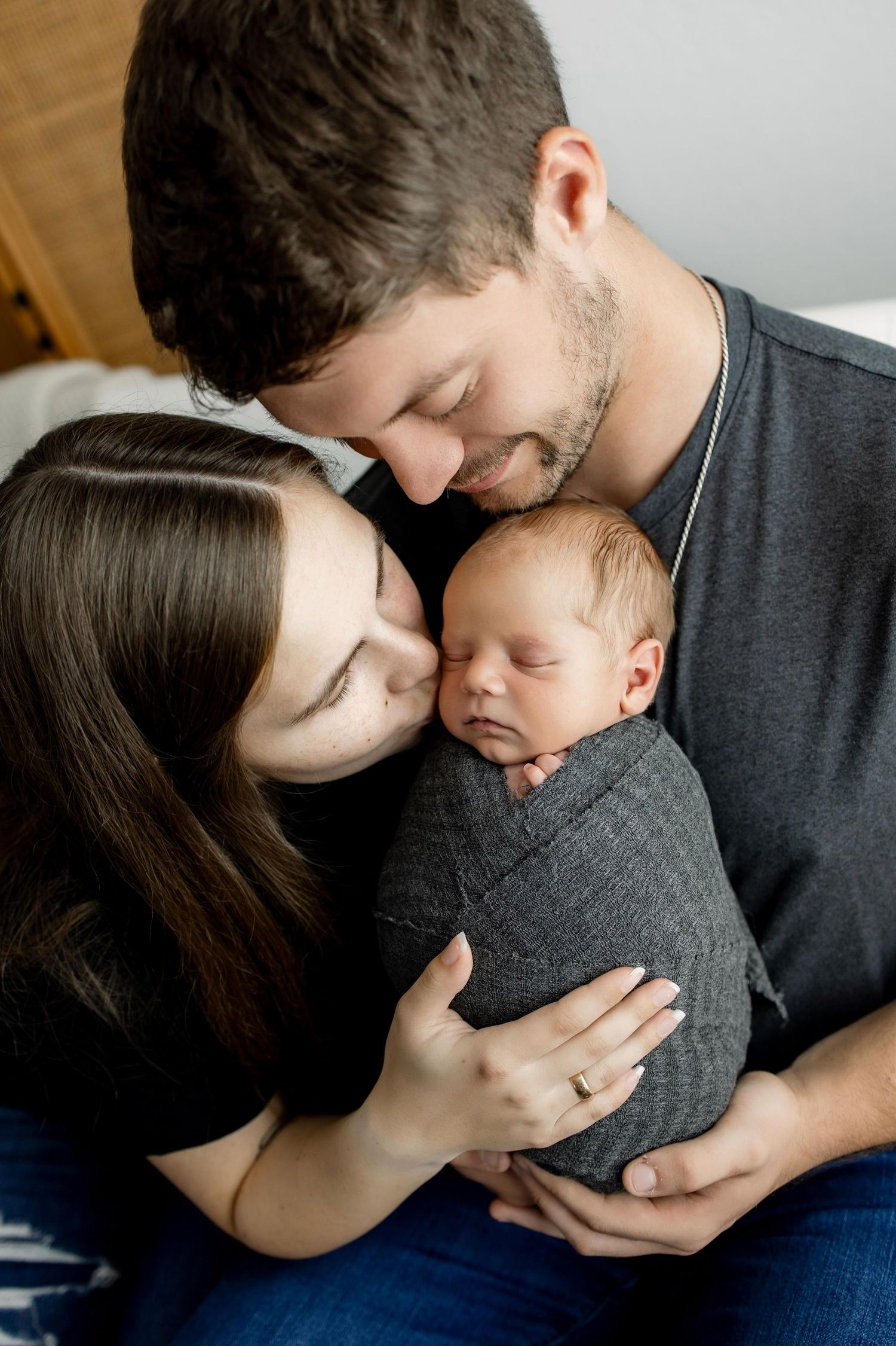 Intimate family portrait of parents cradling newborn baby wrapped in gray swaddle.