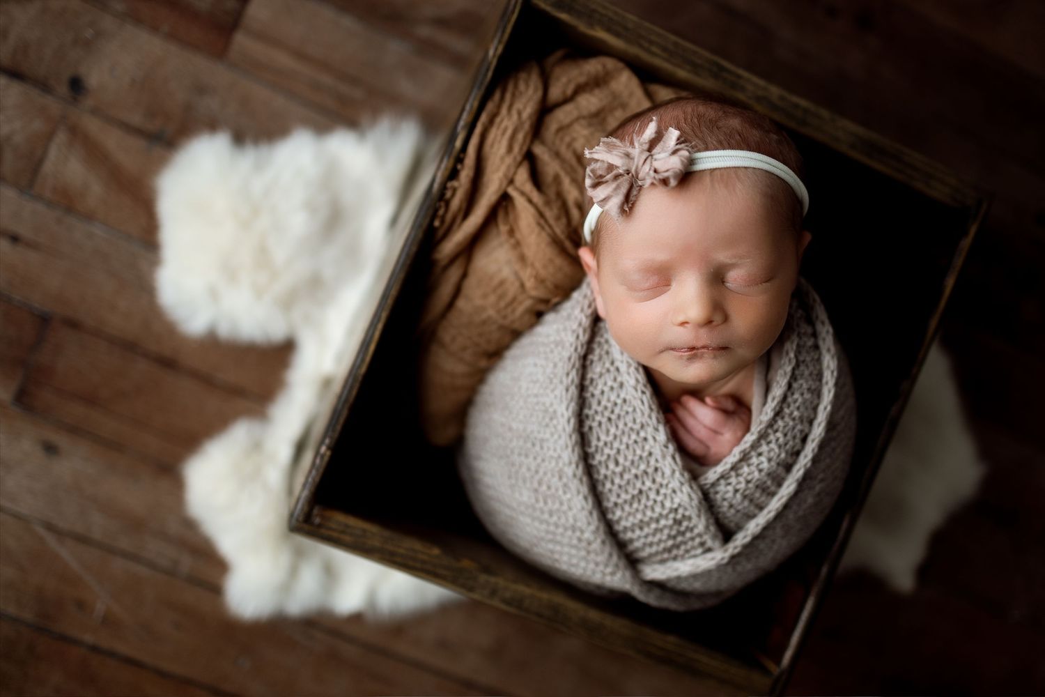 Newborn baby wrapped in gray knit blanket sleeping peacefully in wooden box with white fur accent.