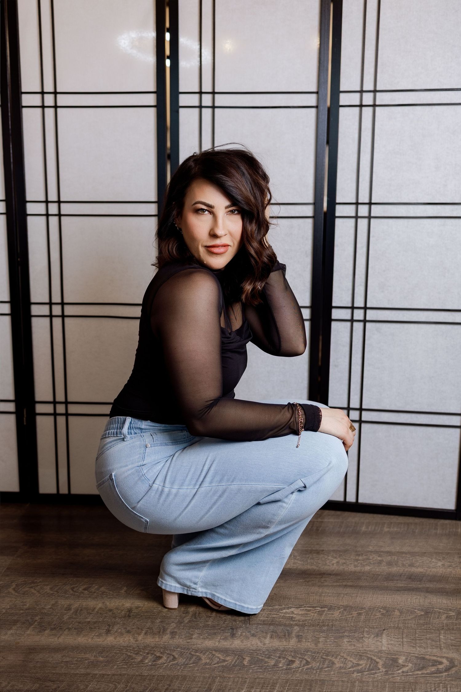 A person in a black top and light jeans poses in a sequence against a white wall with wooden flooring.