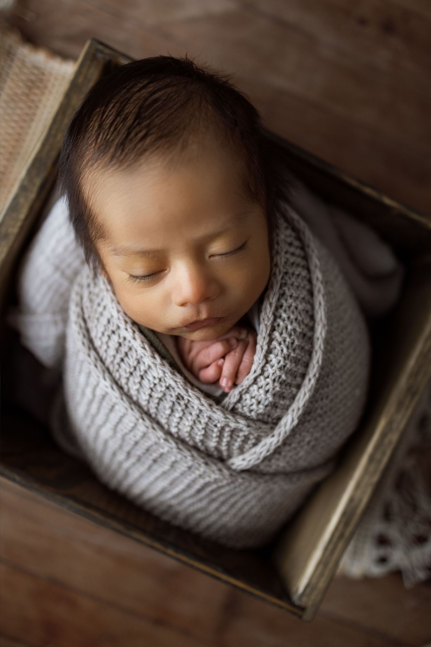 Sleeping newborn baby wrapped in soft gray knit blanket lying peacefully in wooden box from above.