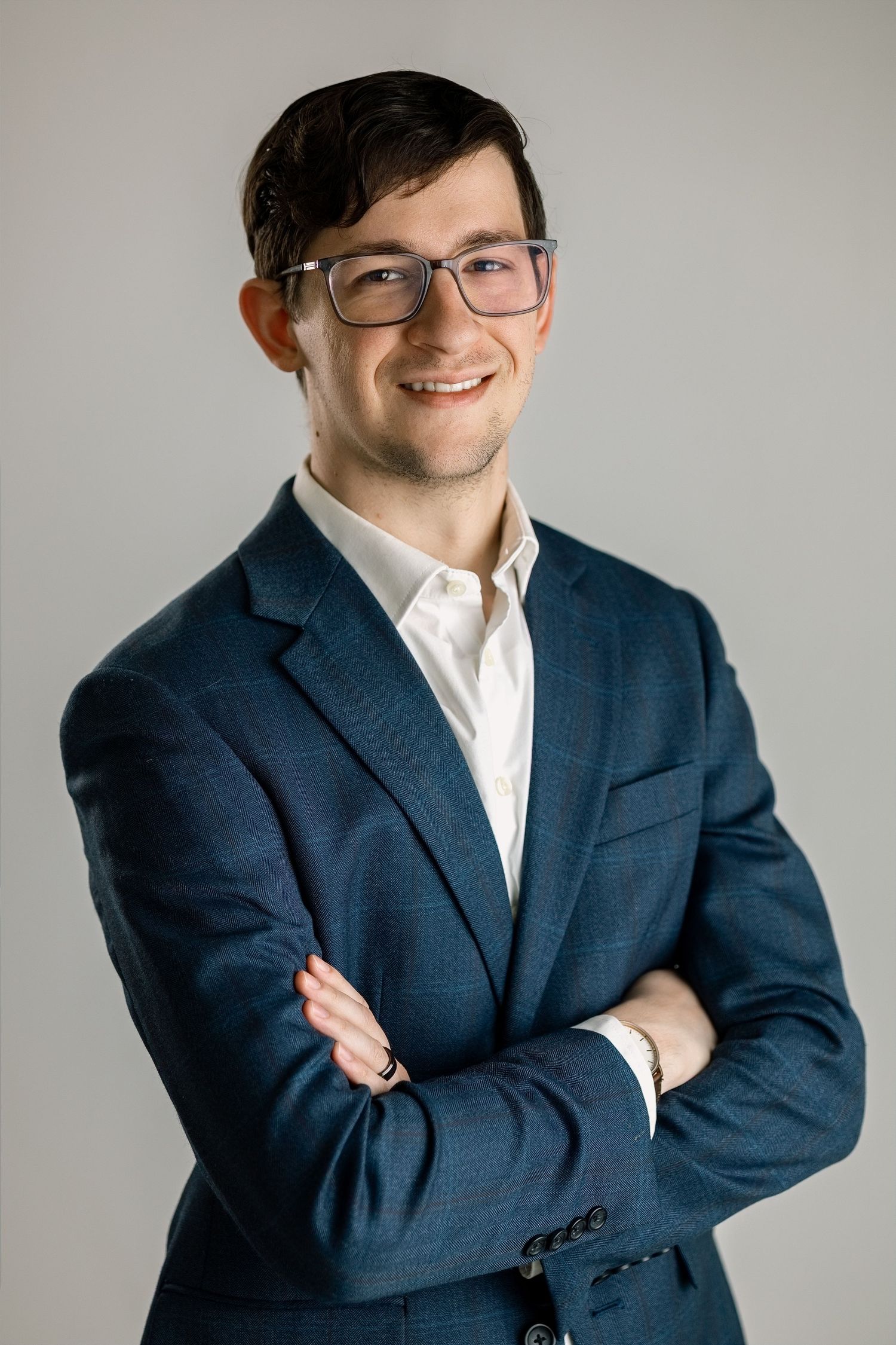 Young professional man in blue suit and glasses smiling with arms crossed against gray background.
