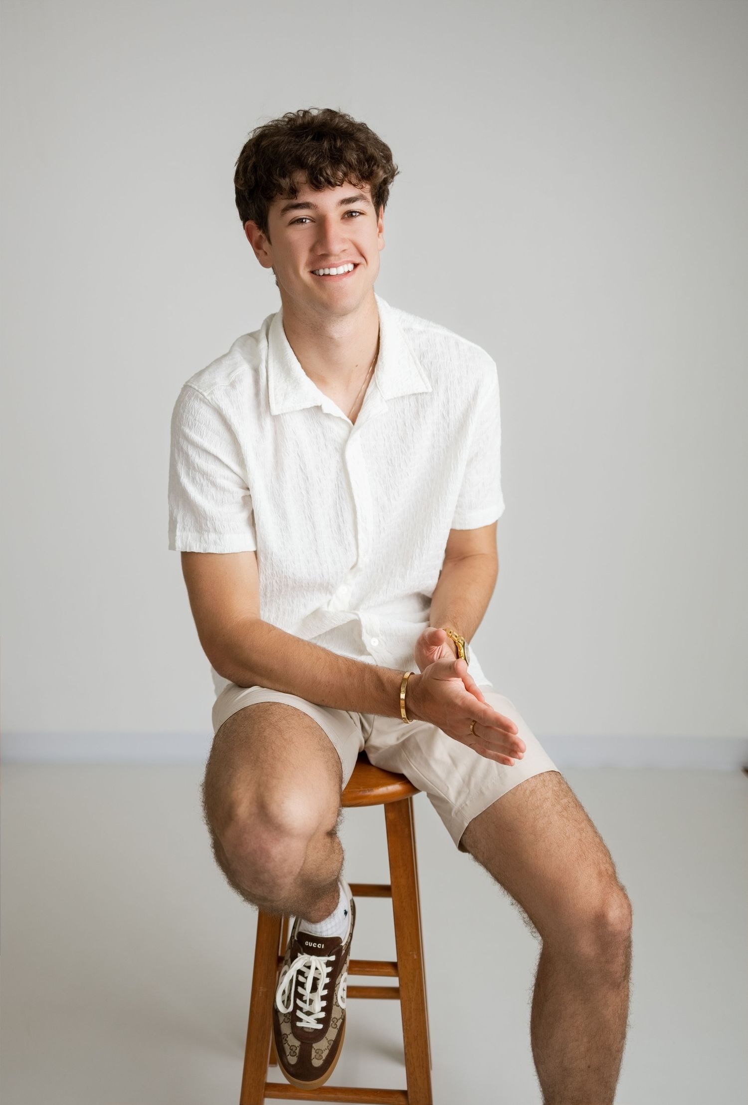 Young man smiling, seated on wooden stool, wearing white short-sleeve shirt and shorts with brown sneakers.