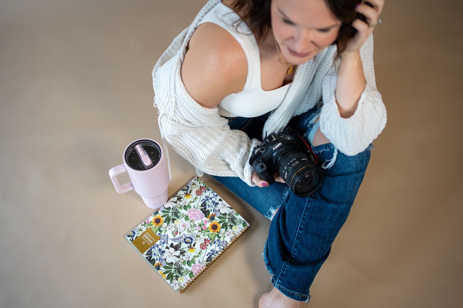 Top down view of someone sitting with a camera, floral notebook and pink coffee cup on beige floor.