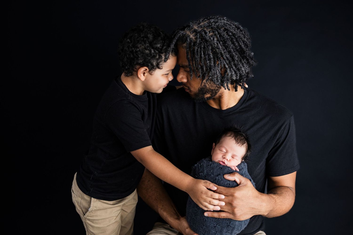 A loving family in matching black shirts embraces while holding a small baby against a dark background.