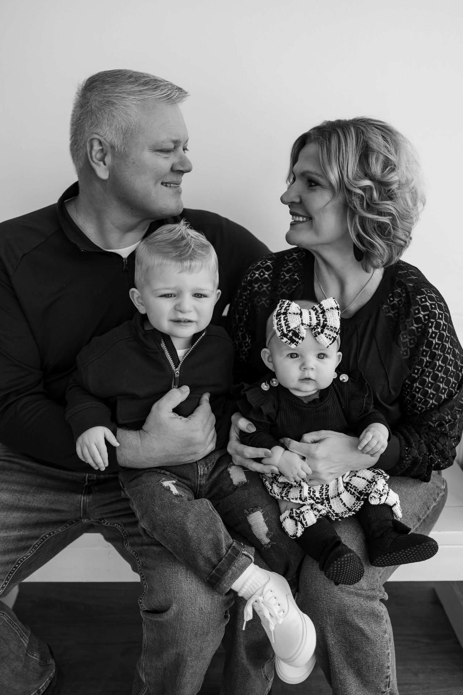 Black and white family portrait of parents smiling at each other while holding two young toddlers on their laps.