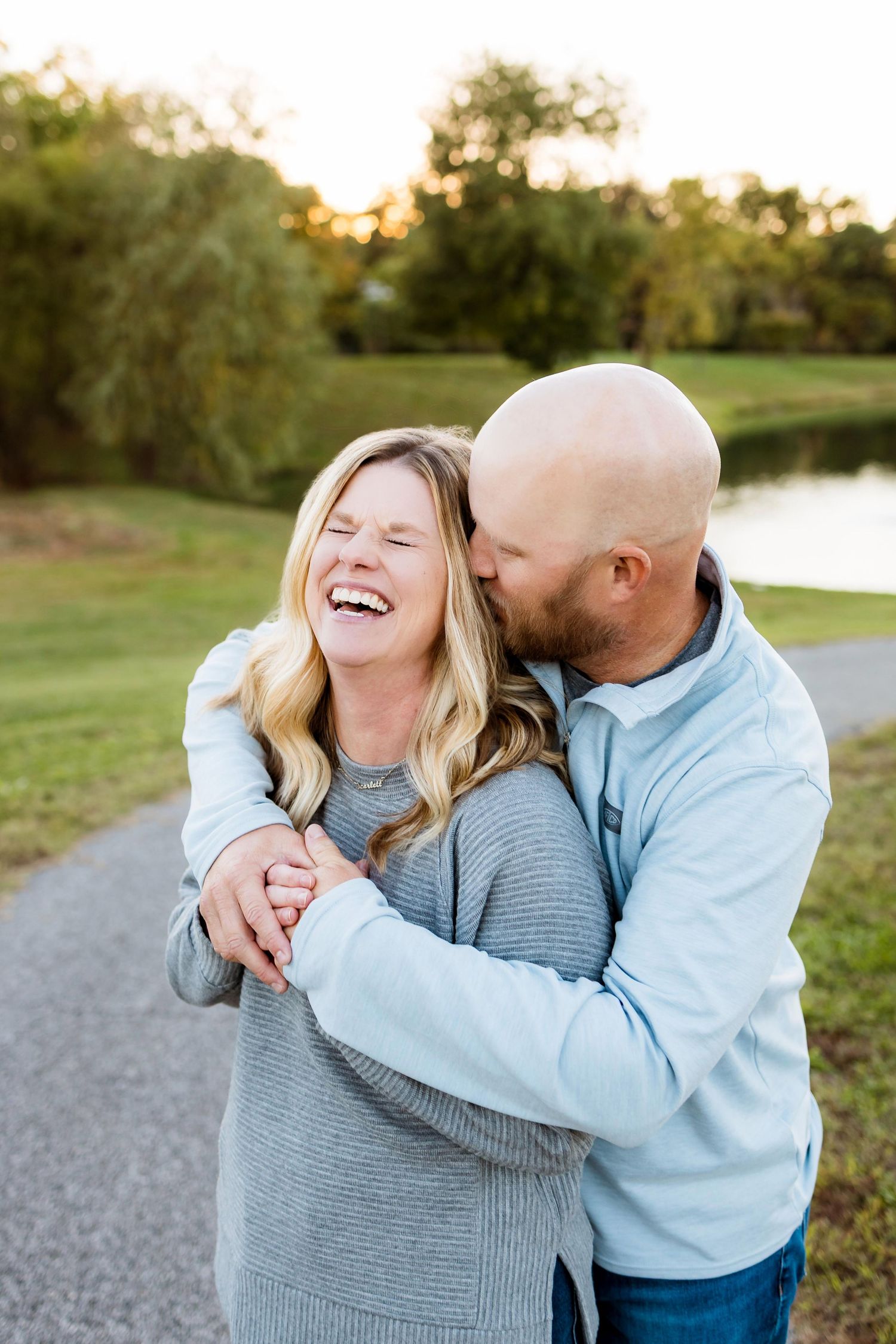A couple shares joyful laughter together on a path during golden hour with trees and a lake in the background.