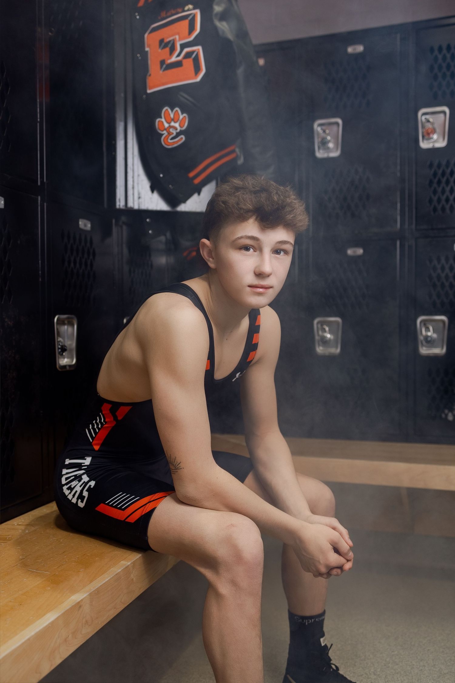 Athlete in black uniform sits thoughtfully on bench in dark locker room setting.
