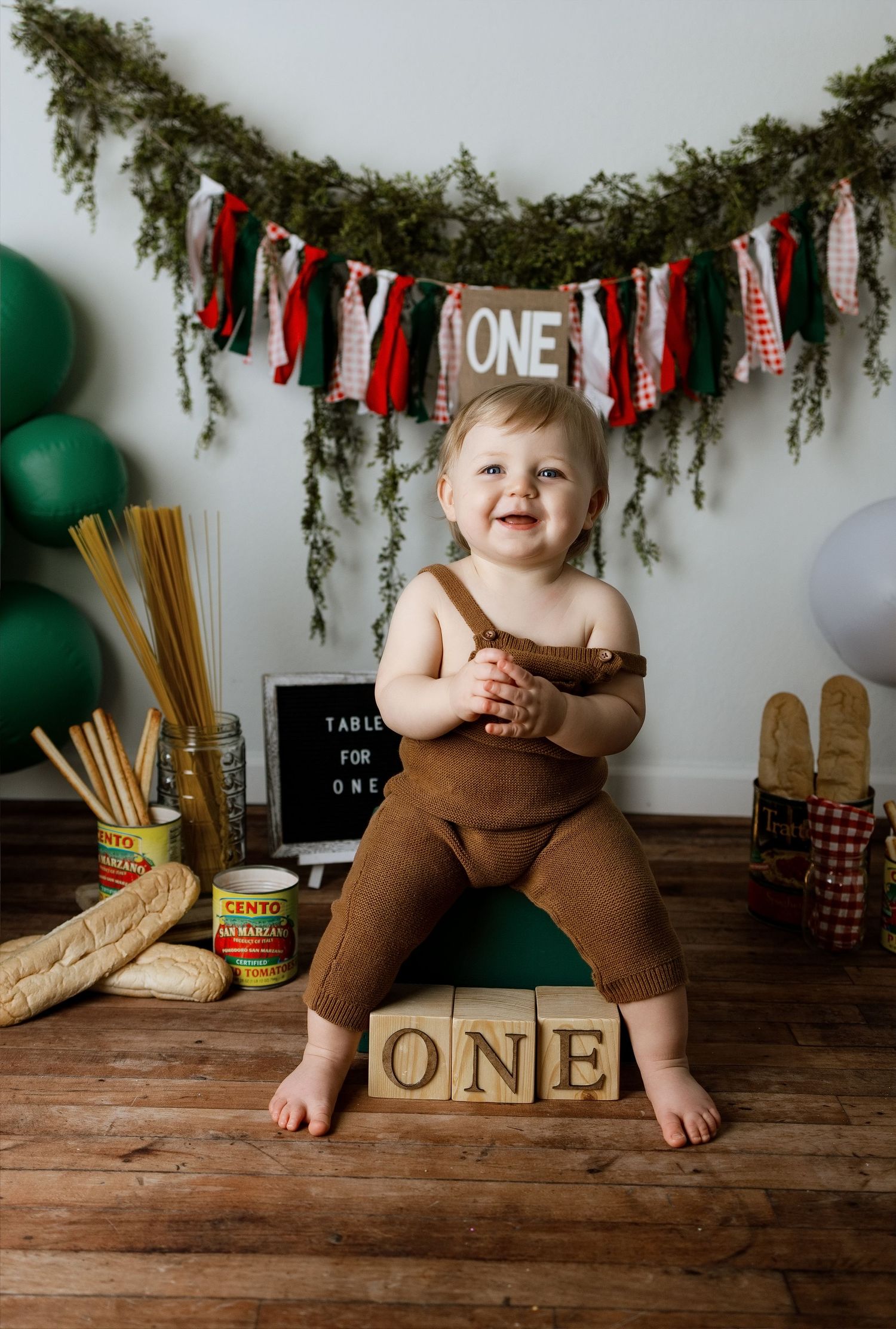 First birthday photo setup with rustic wooden floor, evergreen garland, and red and white banner decorations.