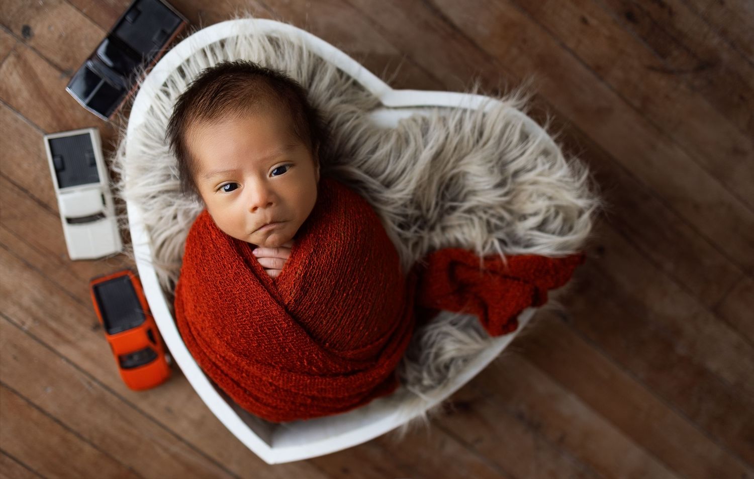 Newborn baby wrapped in red swaddle blanket laying on white fur against wooden floor background.