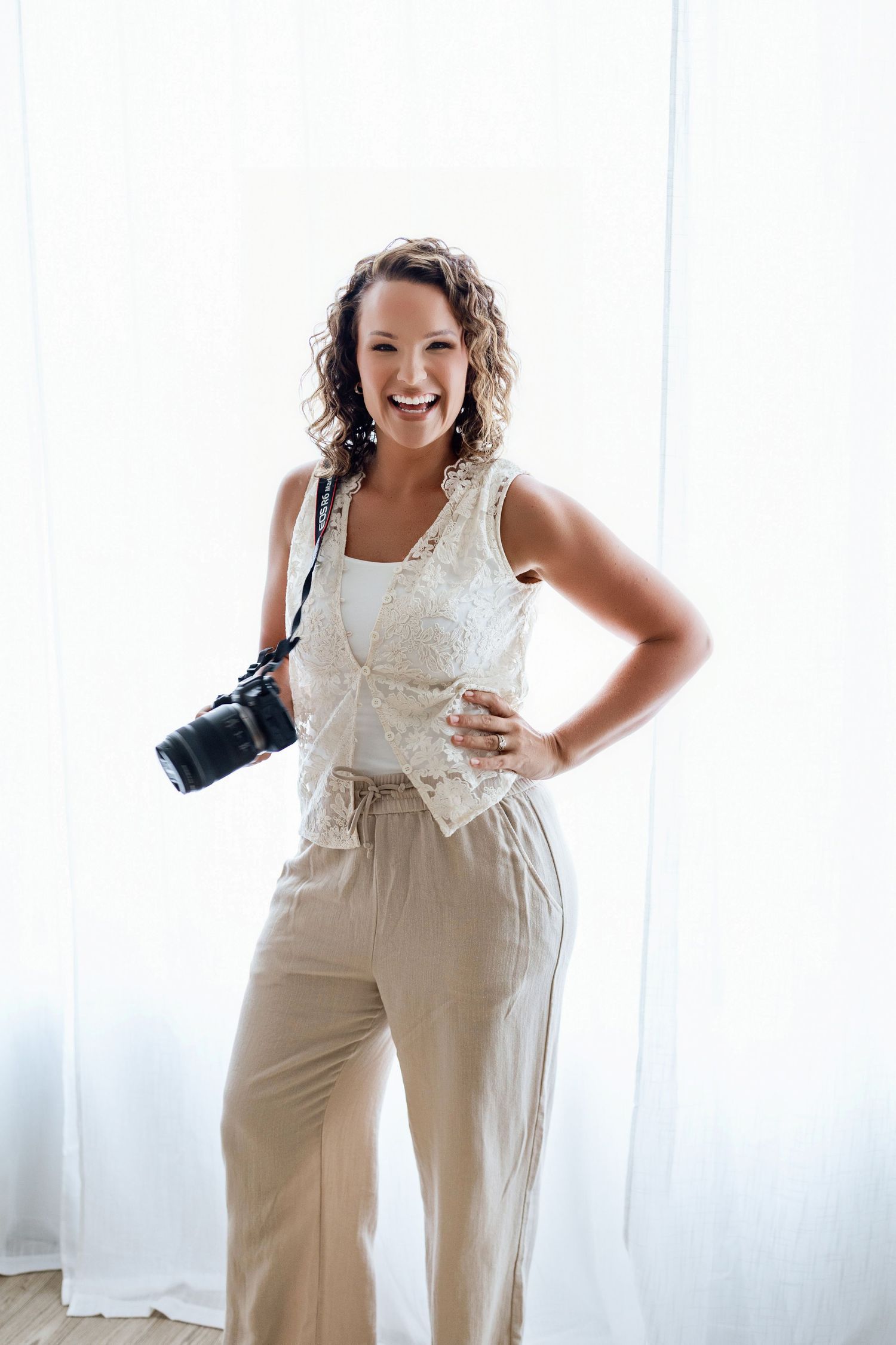 Smiling woman with curly hair holding a camera, wearing beige pants and white lace top, standing in front of white curtains.