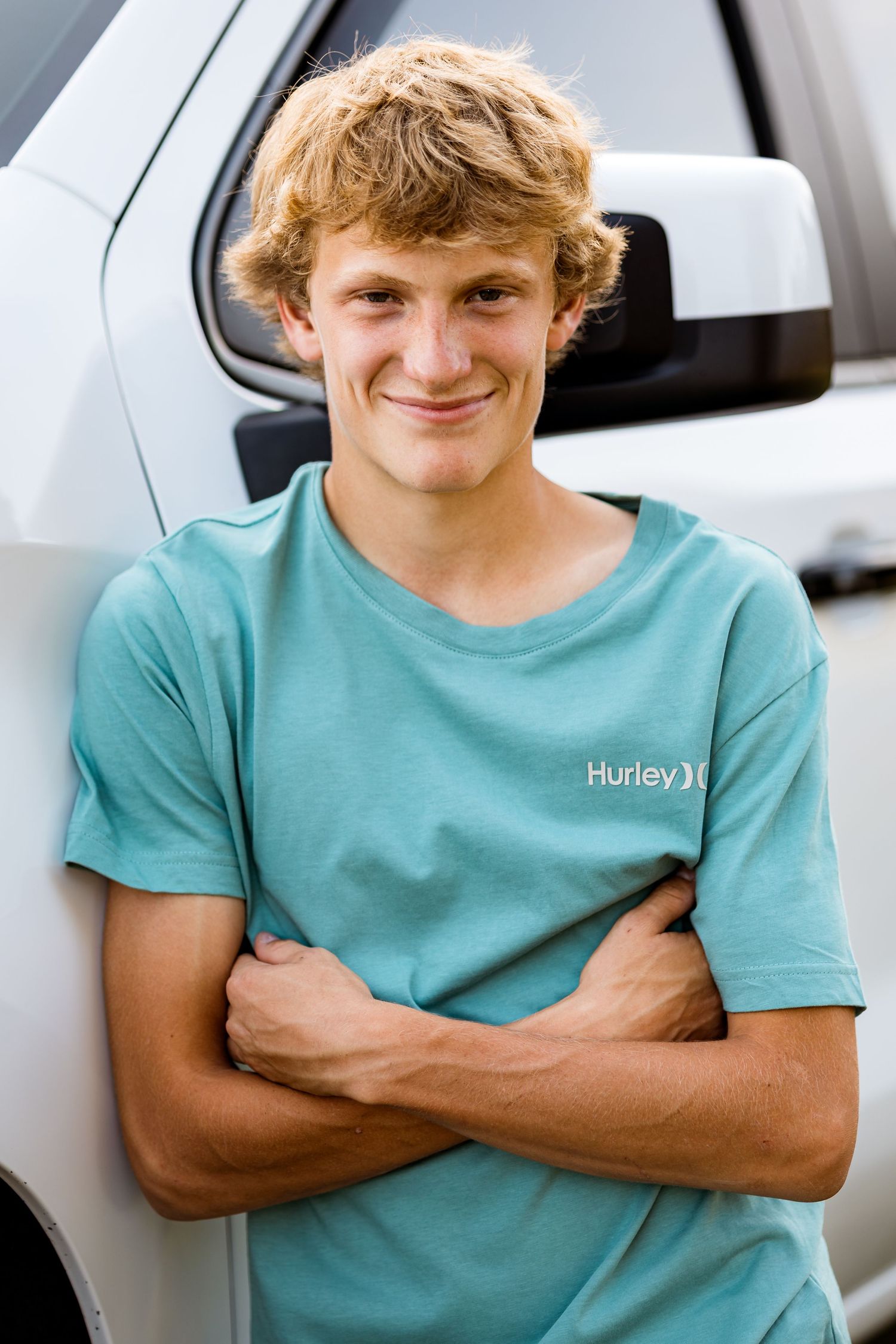 Person in turquoise Hurley t-shirt smiling and posing with crossed arms in front of parked cars.