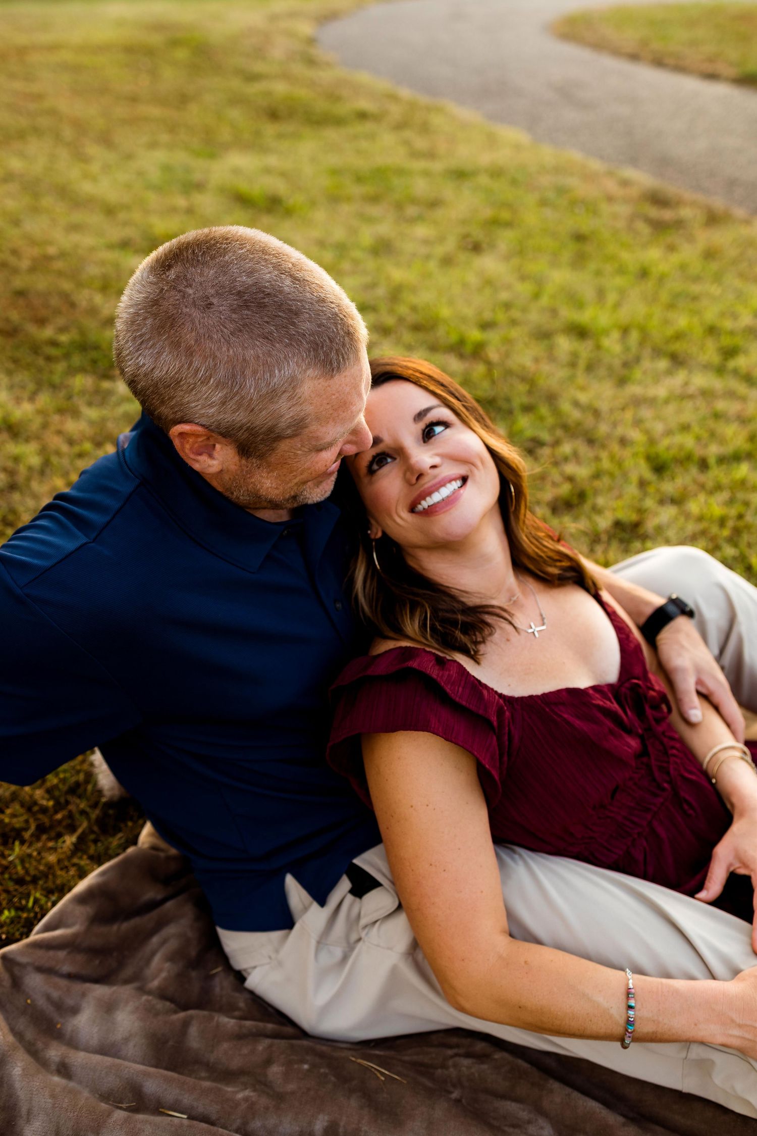 A couple shares an intimate moment while laying on a blanket in a grassy field at sunset.