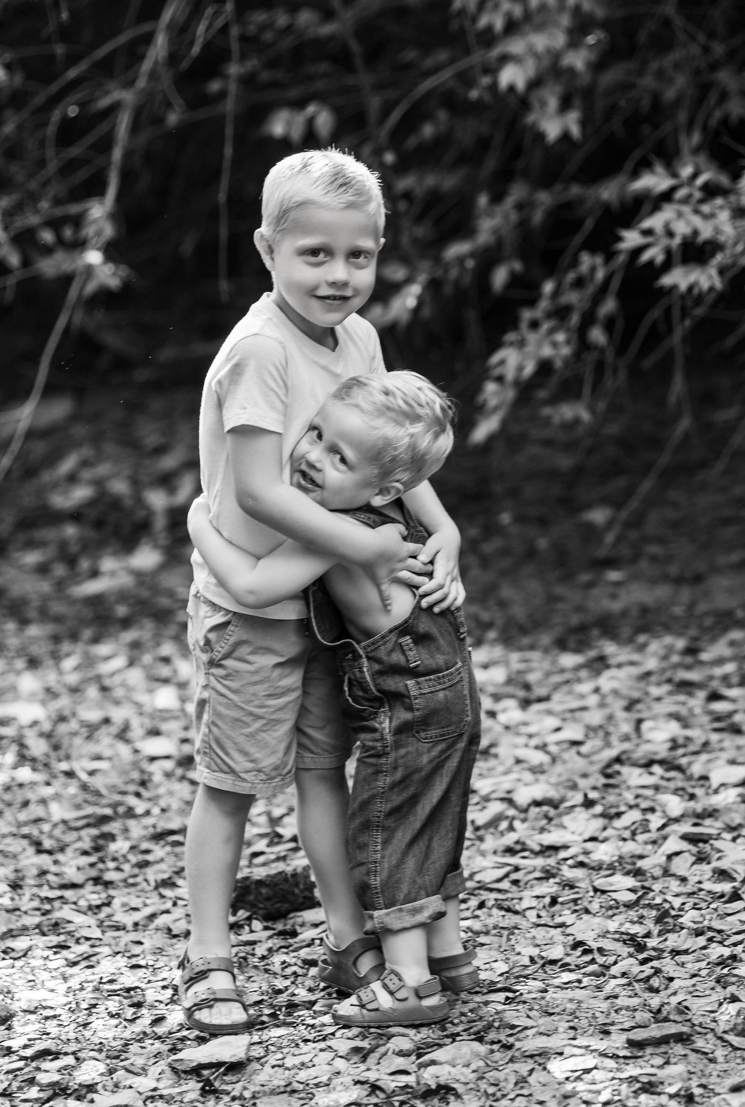 Two young children in overalls hug and play together on a path covered in fallen leaves in black and white.