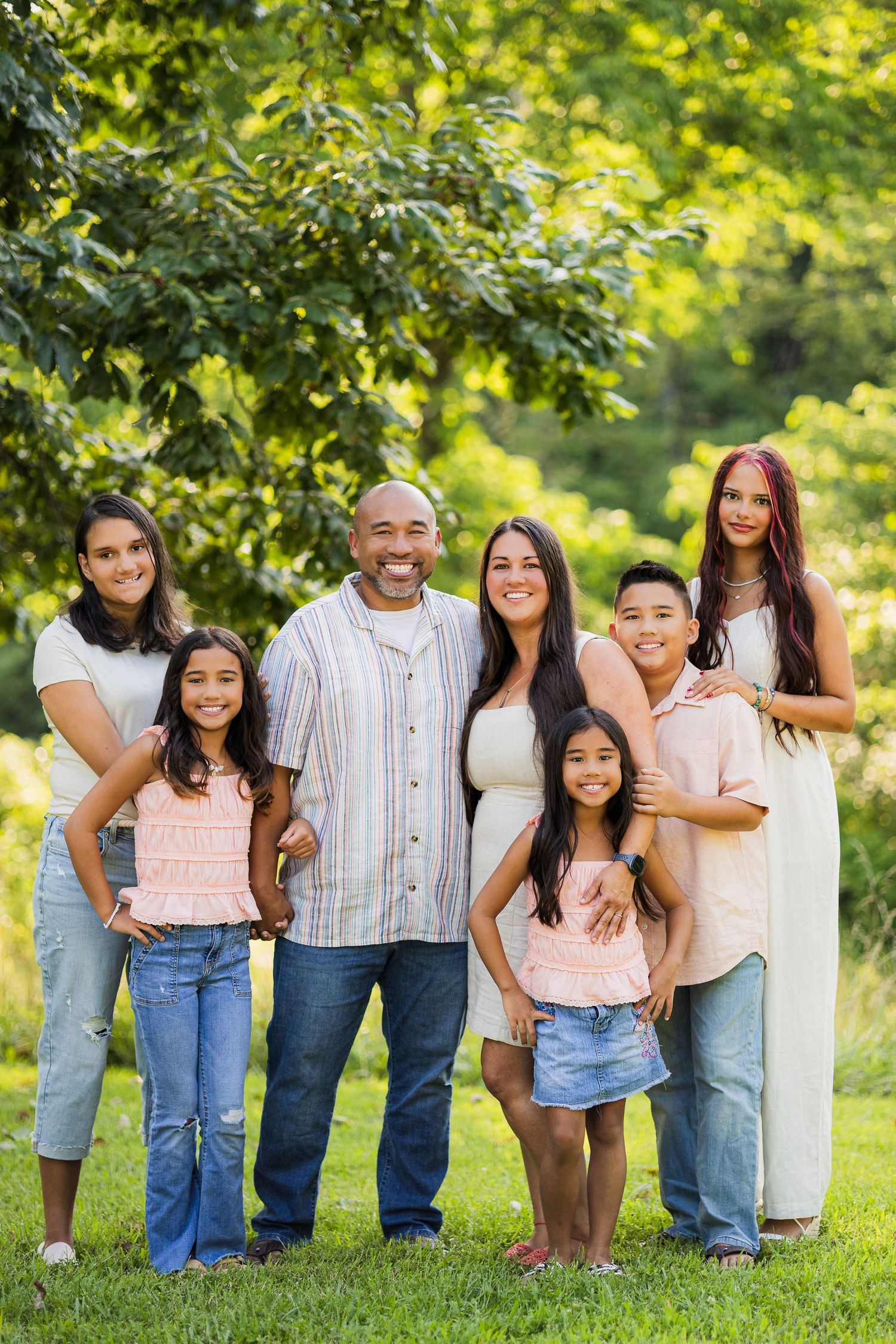 A happy family poses together outdoors on a sunny day with lush green trees in the background.