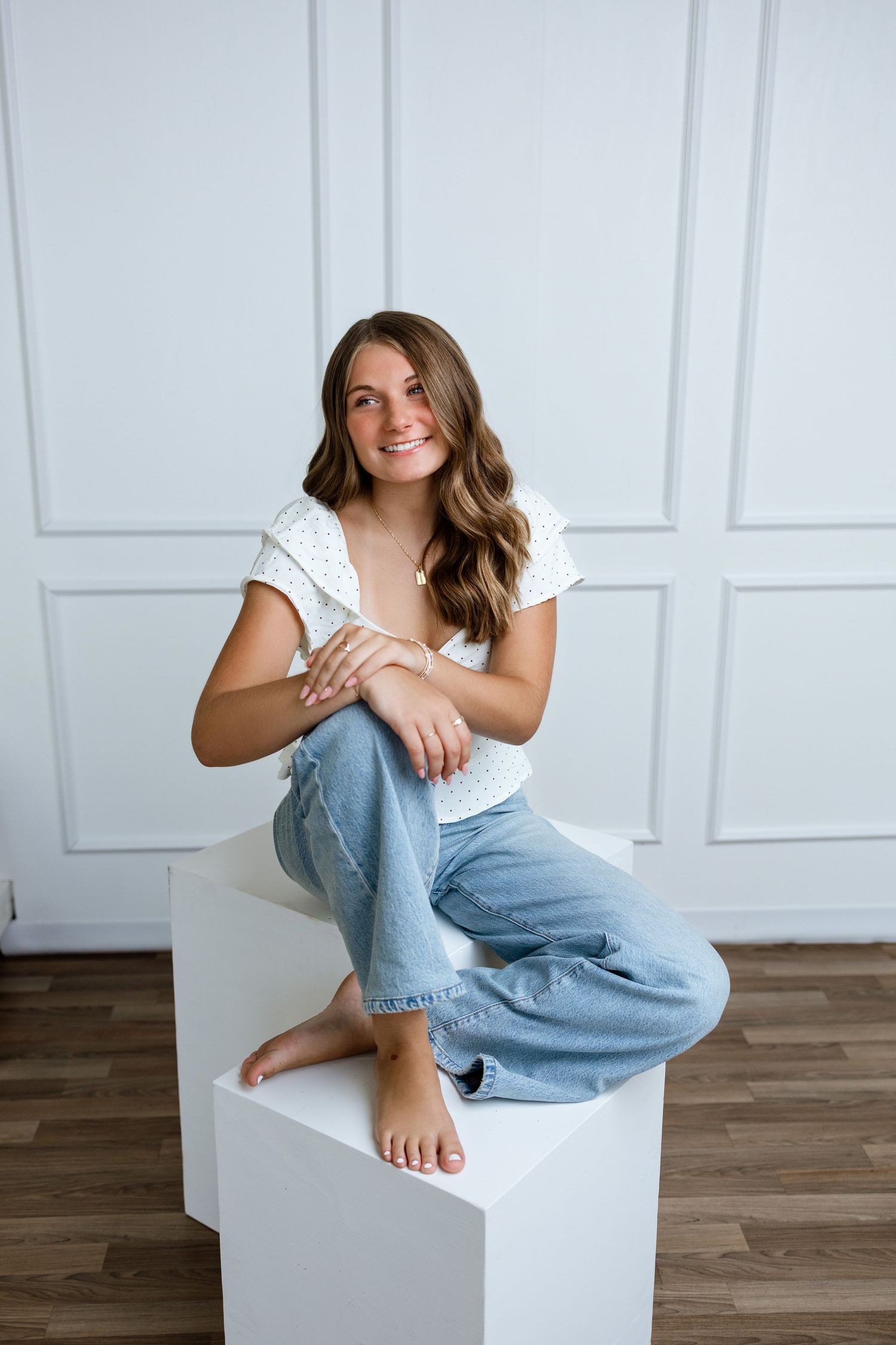 Young woman in white top and blue jeans sitting barefoot on white cube against white paneled wall.