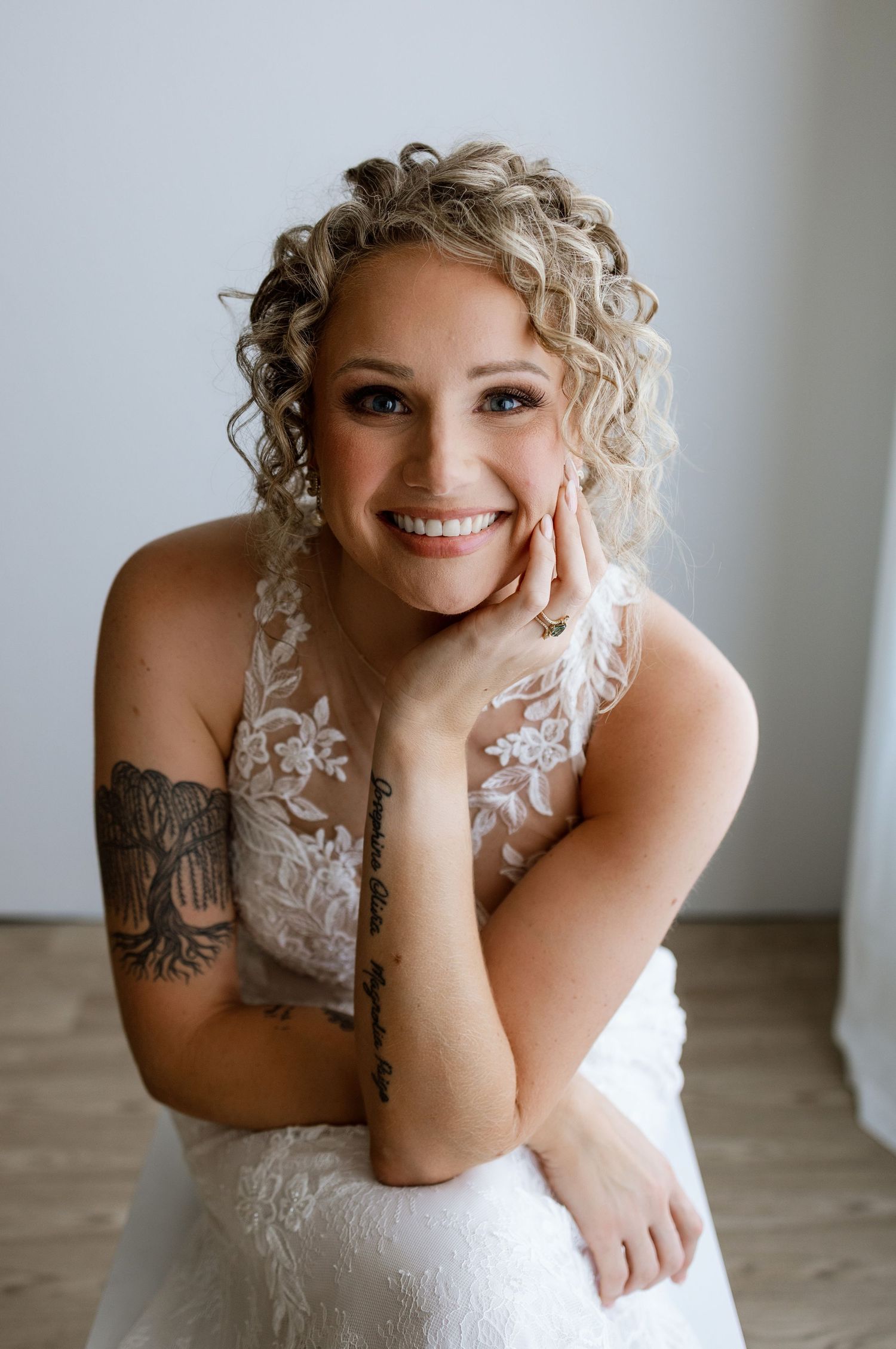 Smiling bride in lace wedding dress with curly blonde hair and tattoos poses gracefully for bridal portrait.