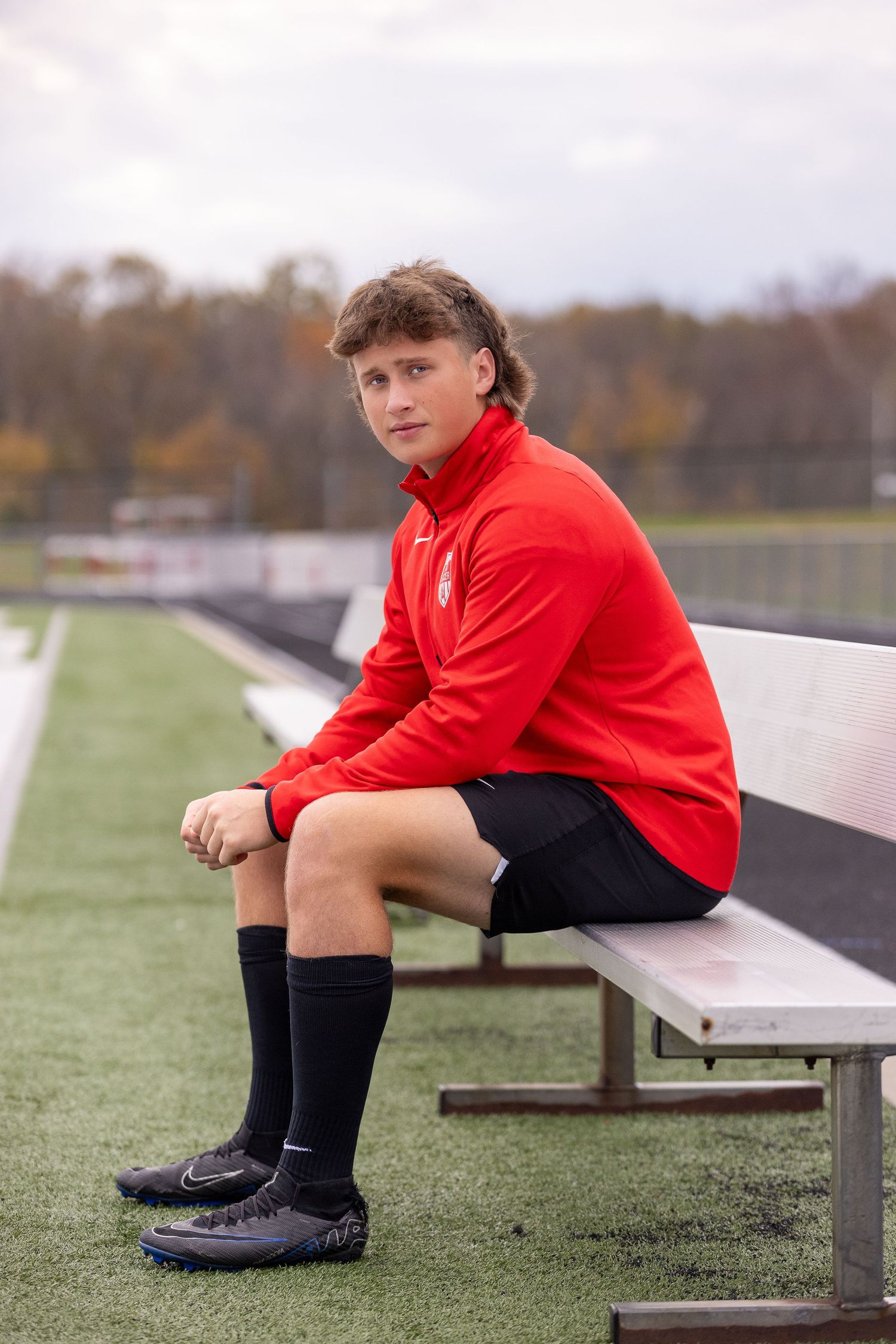 Athlete in red jacket and black shorts sitting on bleachers at a sports field.