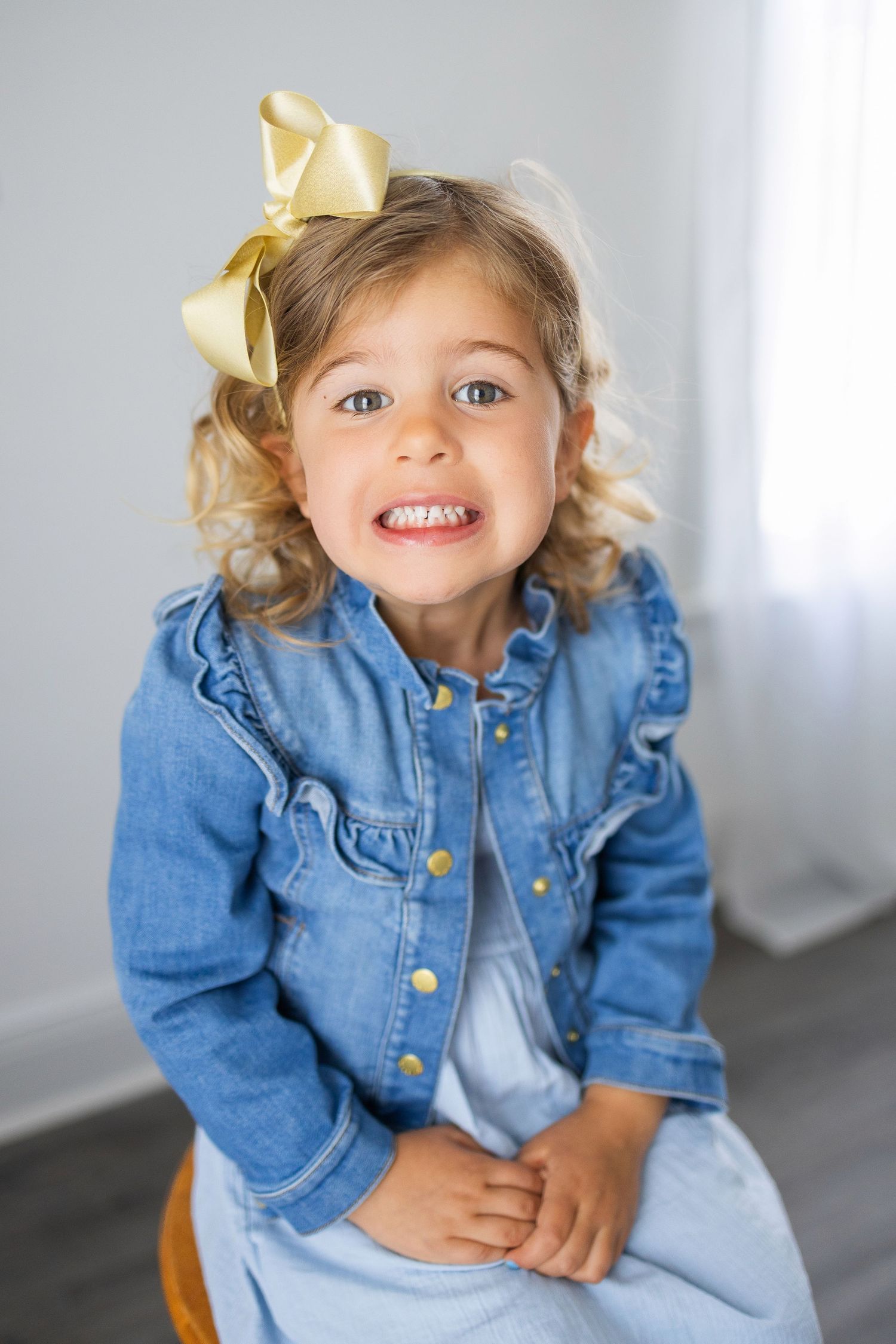 Smiling child wearing a denim jacket and yellow hair bow sits casually on gray chair.