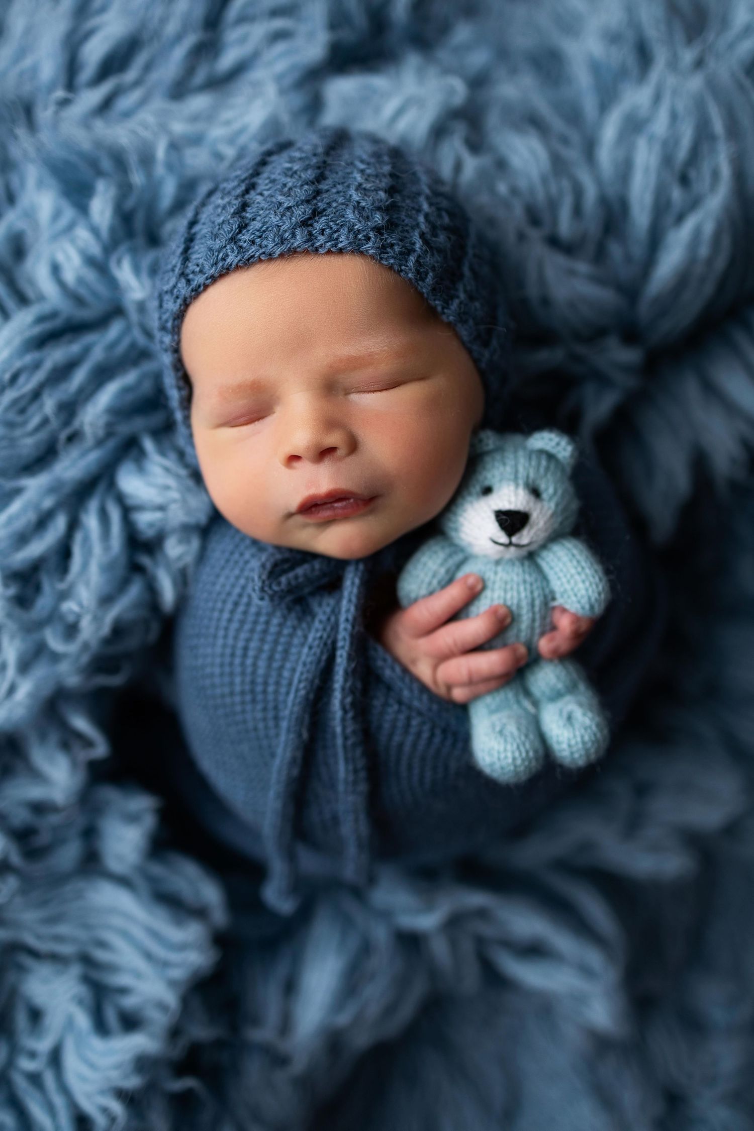 Sleeping newborn in blue knit wrap cuddling with small light blue teddy bear on textured blanket.