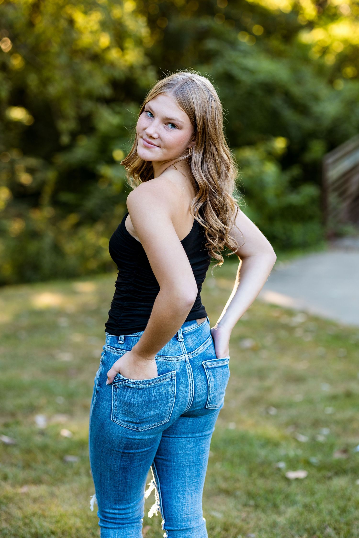A person in a black tank top and blue jeans poses outdoors on a sunny day with greenery in the background.