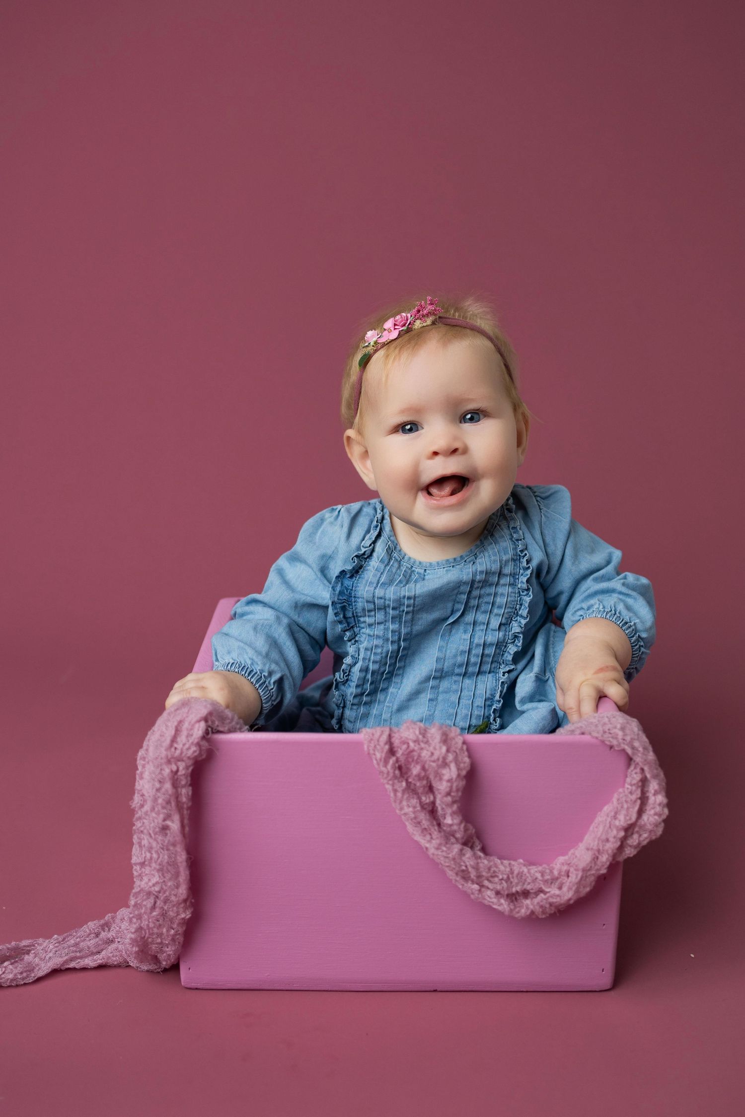 Baby in blue dress poses inside pink photography prop box against mauve backdrop in professional studio setting.