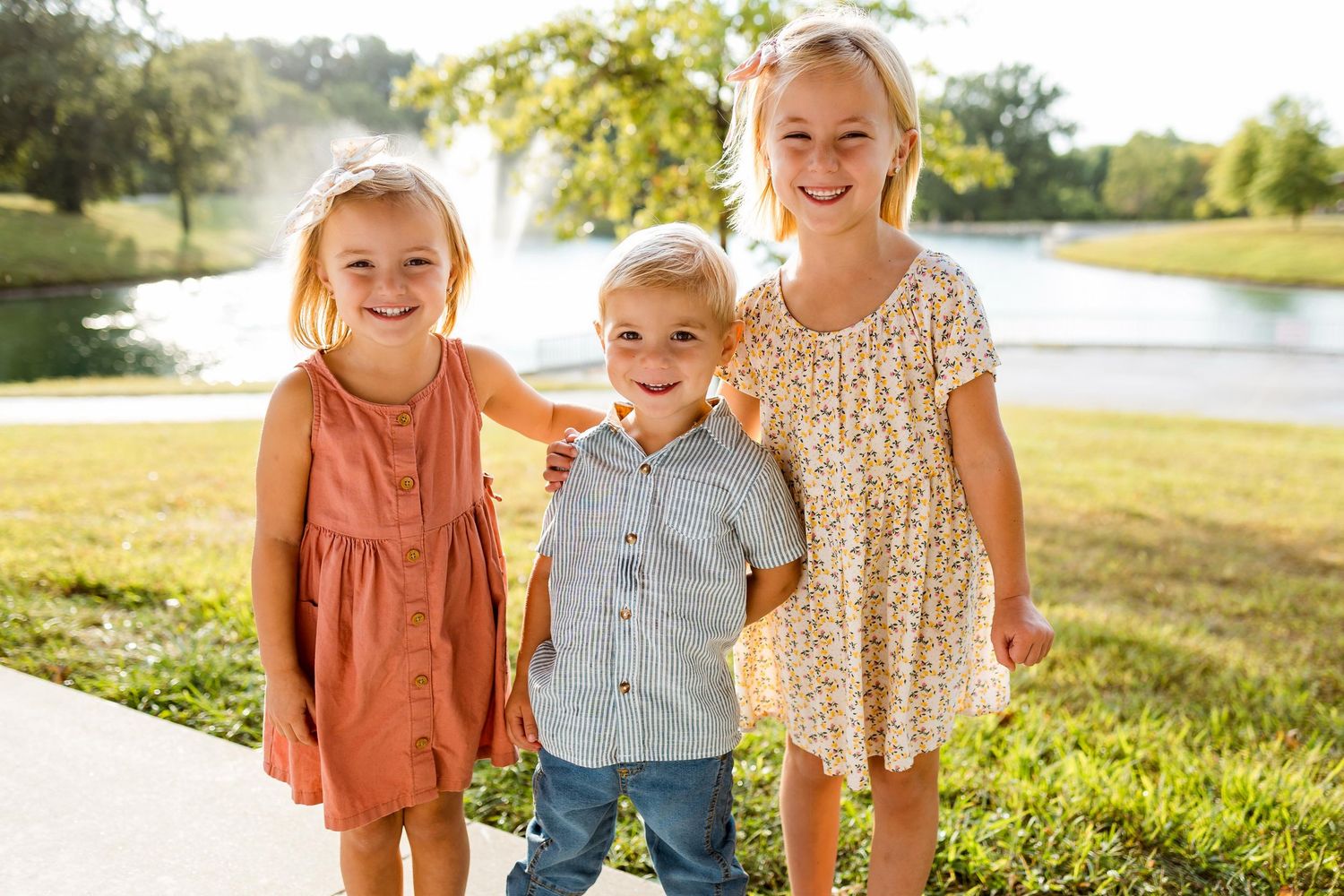 Three children stand together outdoors in casual clothing on a sunny day in a grassy field.