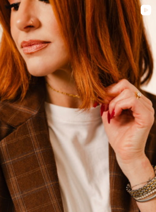 A woman with vibrant red hair wearing a brown plaid blazer, white tee, gold necklace, and silver bracelets.