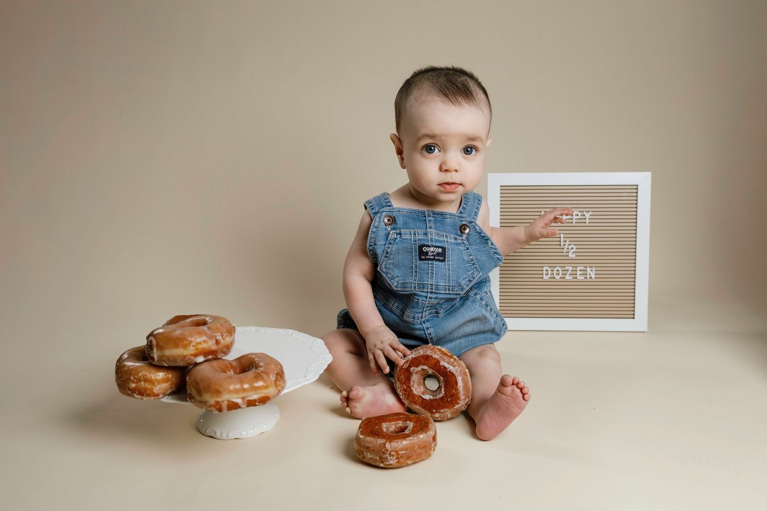 A baby in denim overalls sits on a beige backdrop with fresh baked cinnamon rolls arranged around them.