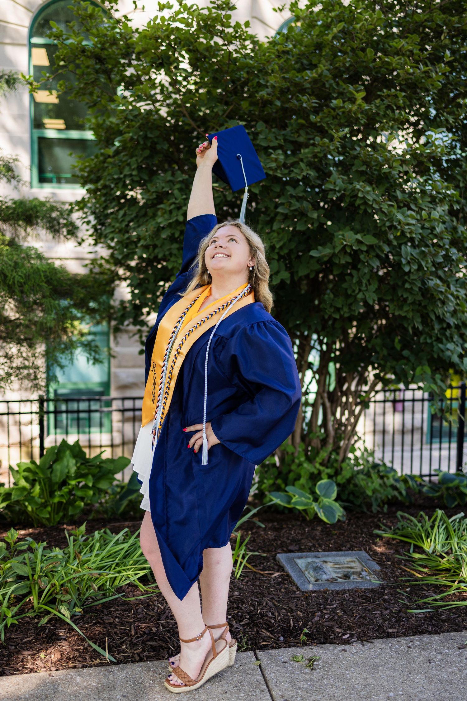 Graduate in blue cap and gown celebrates with joyful pose outdoors on campus while wearing gold cords.