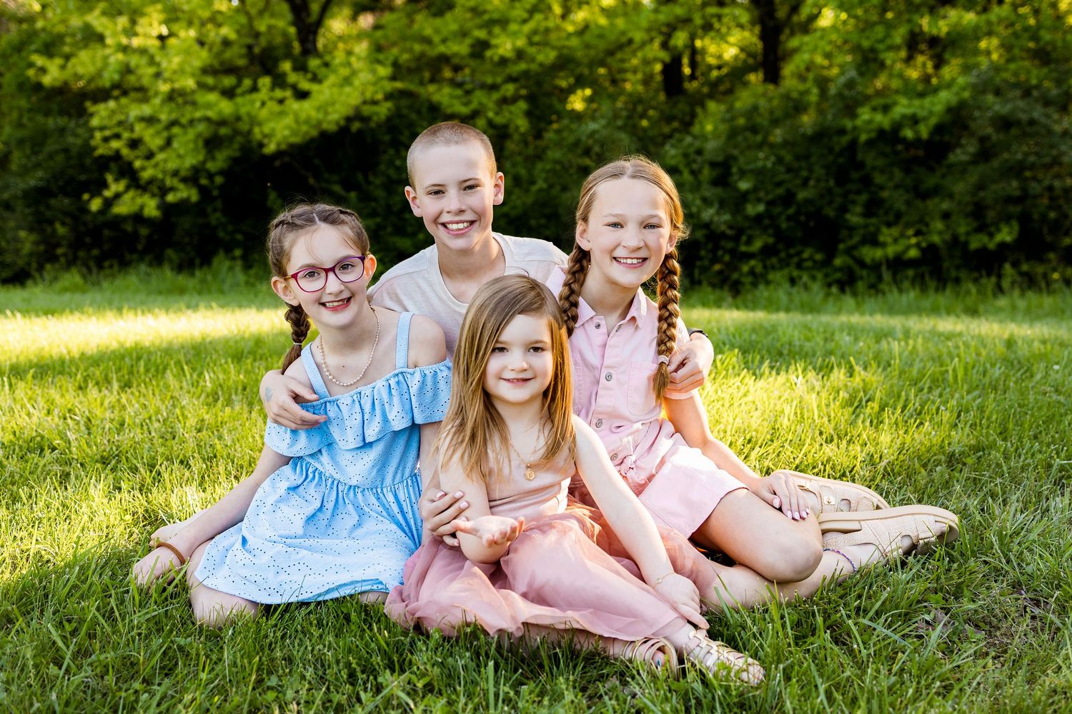 Four children sitting together on blanket in sunny grassy field during summer photoshoot.