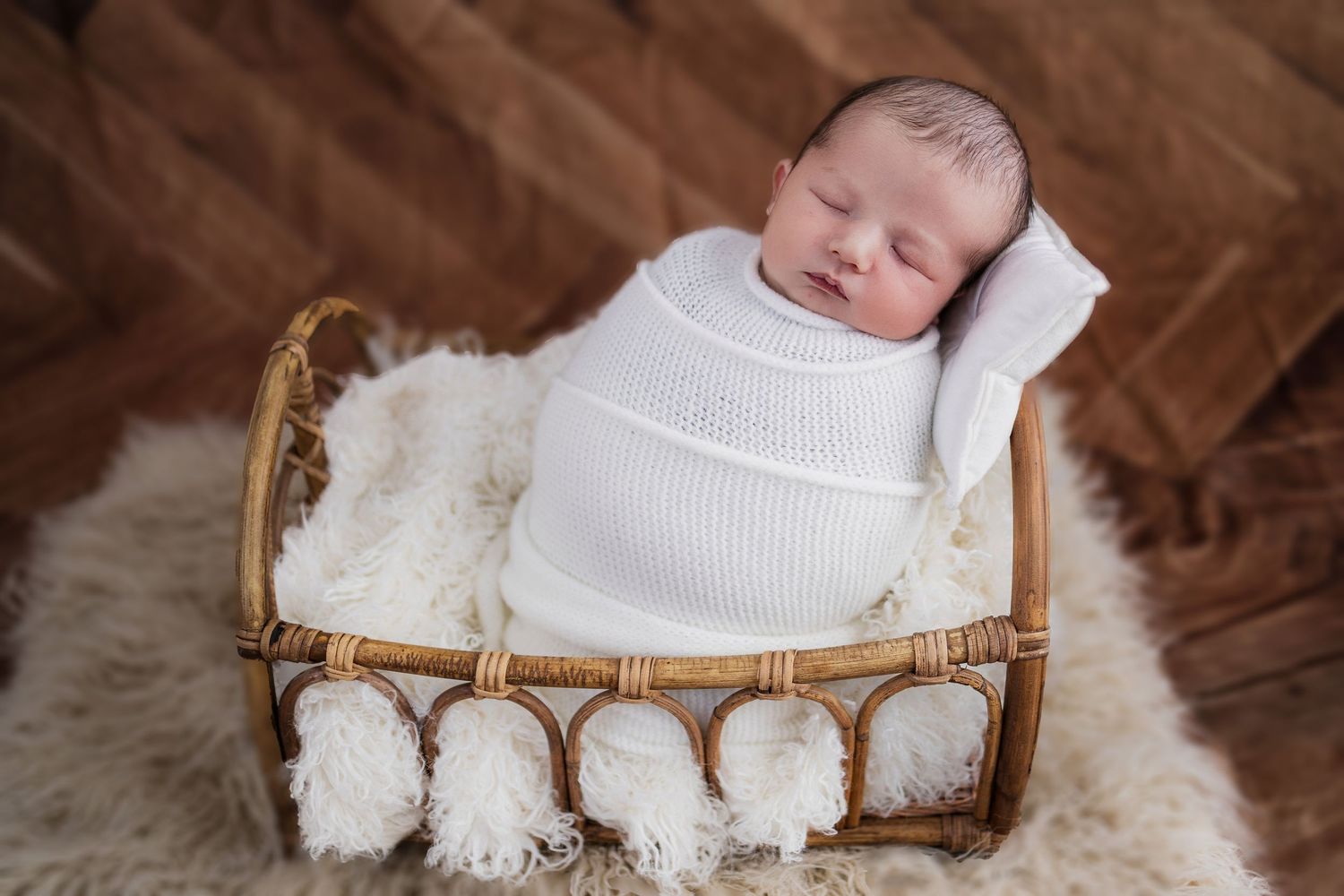 A sleeping infant wrapped in white swaddling lies in a bamboo basket lined with plush white fur.