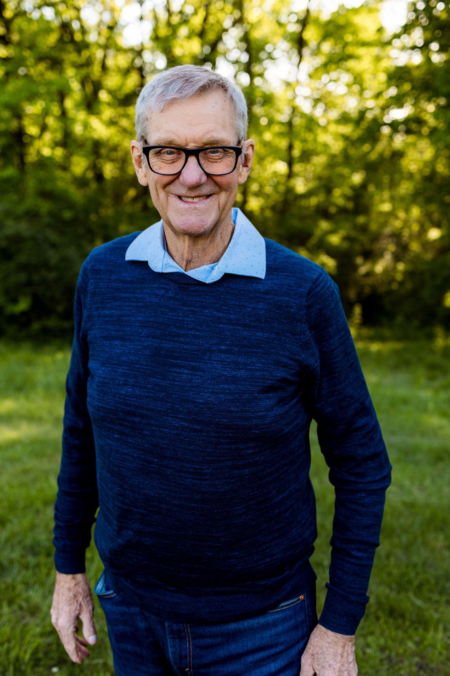 Senior man in navy sweater and glasses smiles warmly against lush green background.