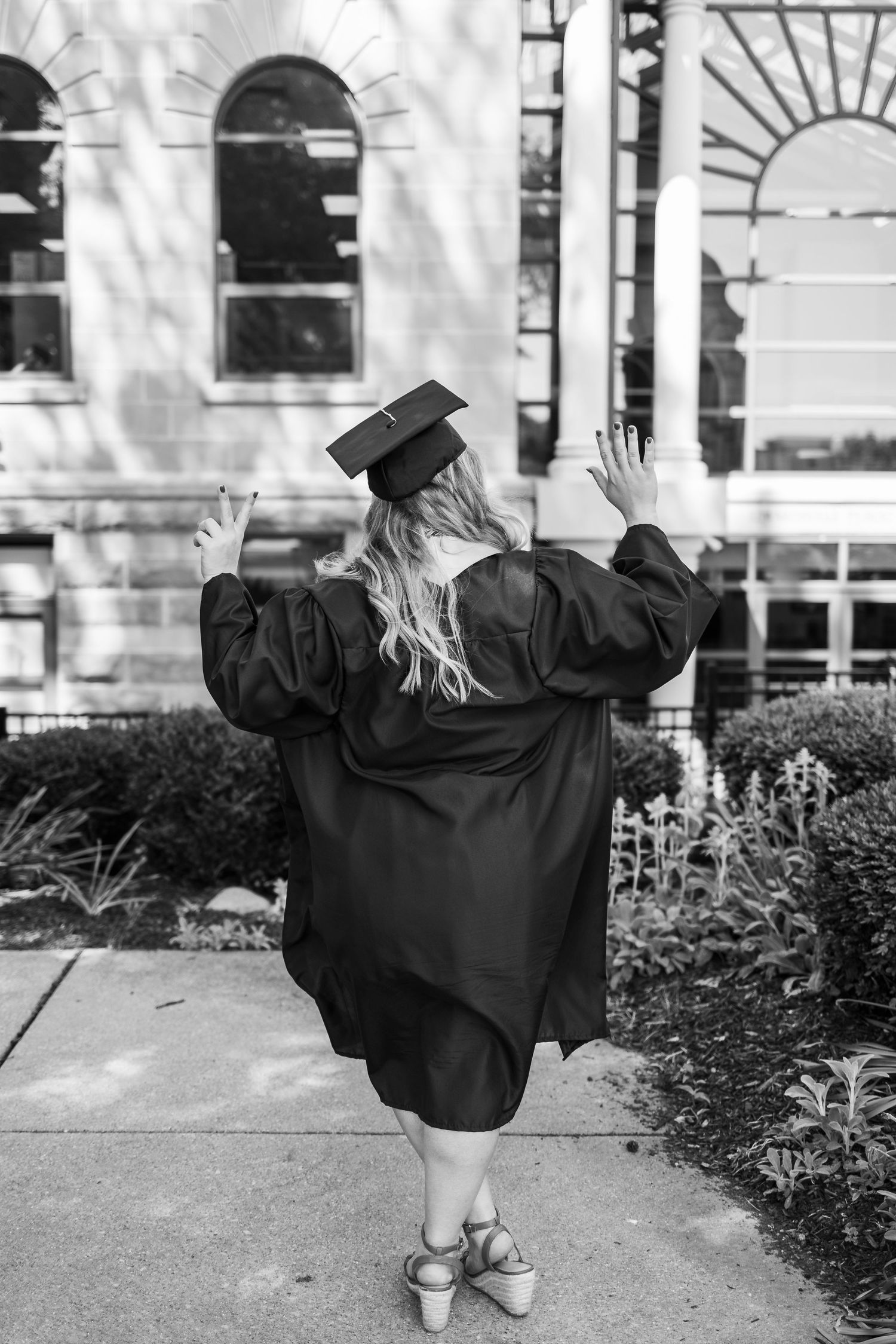 Graduate in black cap and gown raises arms in celebration outside historic university building.