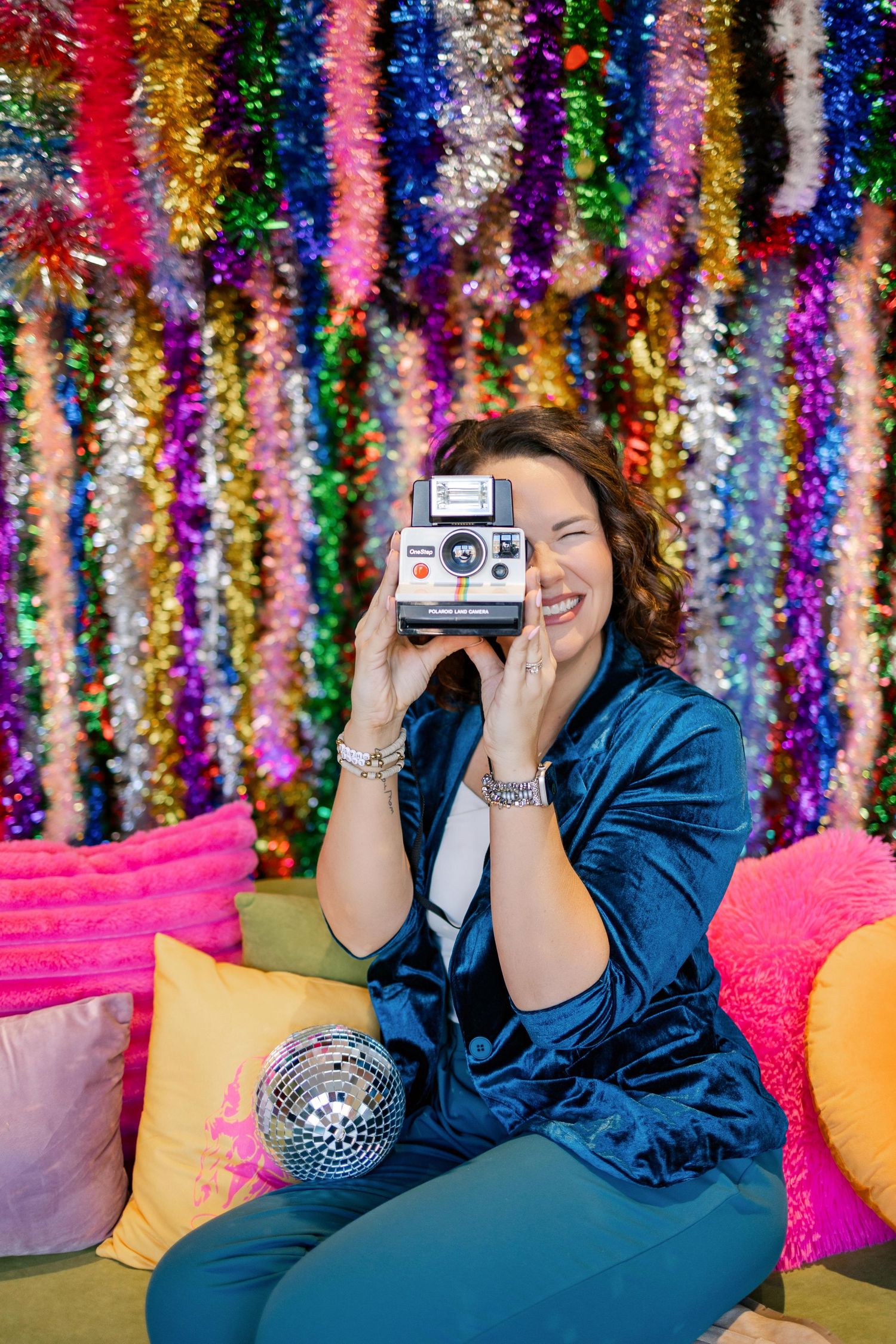 Person taking photo with vintage Polaroid camera against colorful tinsel backdrop on cozy couch with pillows.