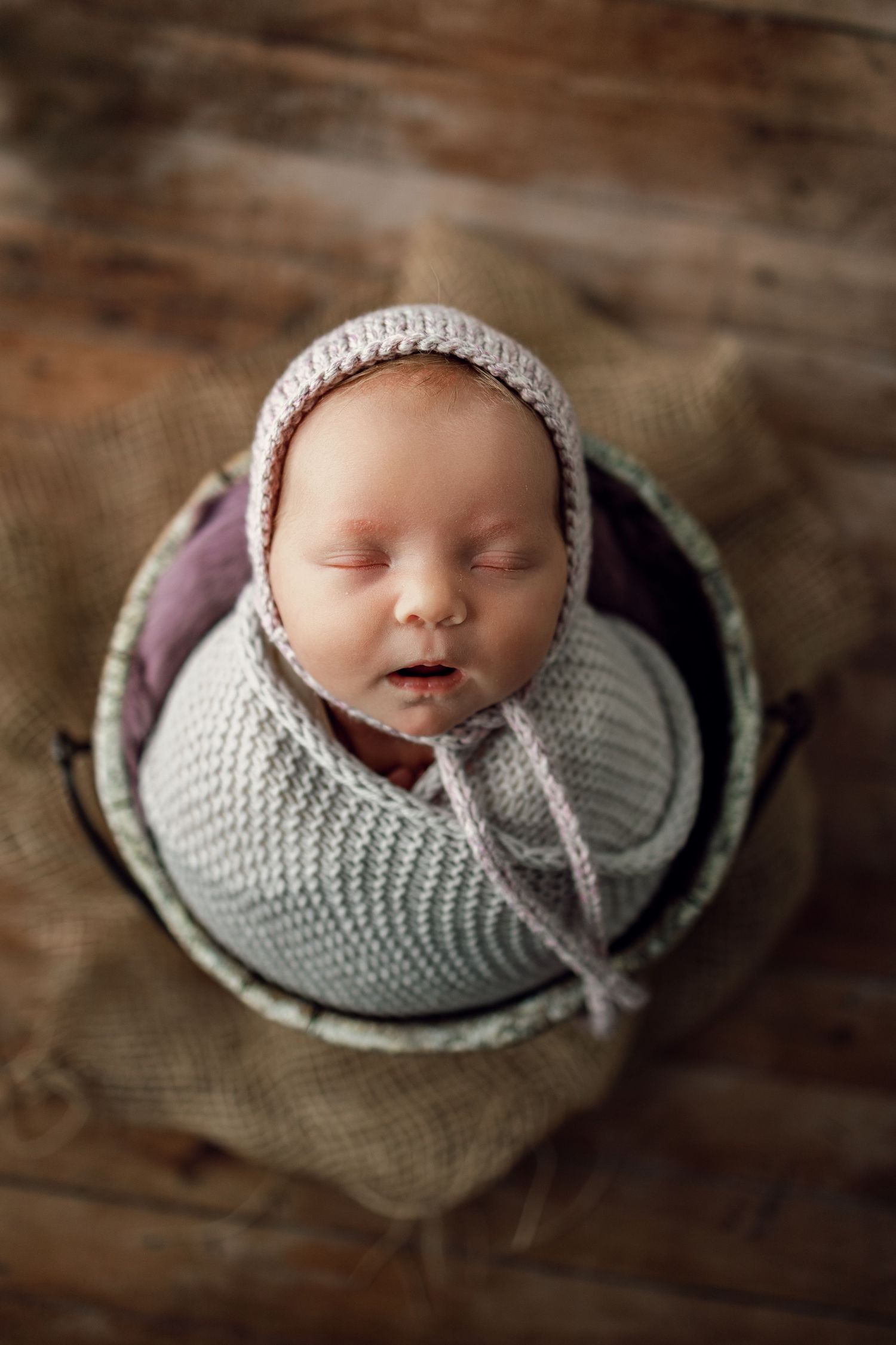 A sleeping newborn baby swaddled in a soft gray knit wrap on a rustic wooden surface.