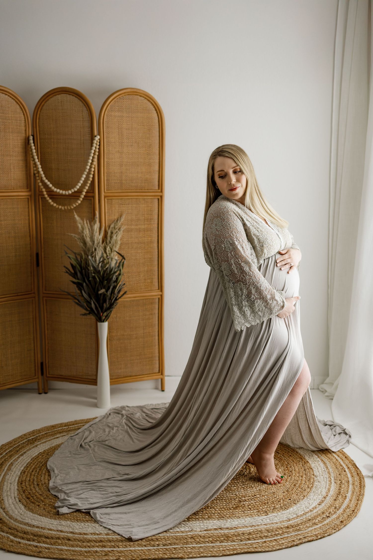 Maternity photoshoot featuring a flowing grey dress with lace details against a minimalist backdrop with woven rug.