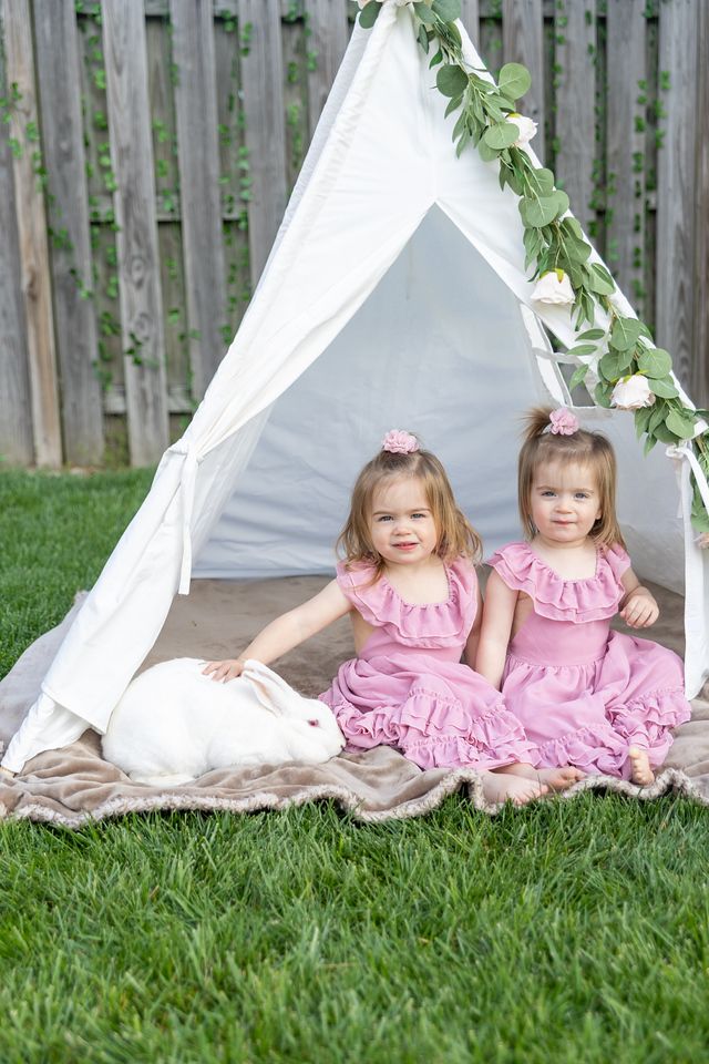 Two young children in matching pink dresses sit together by white teepee tents decorated with greenery in a backyard.