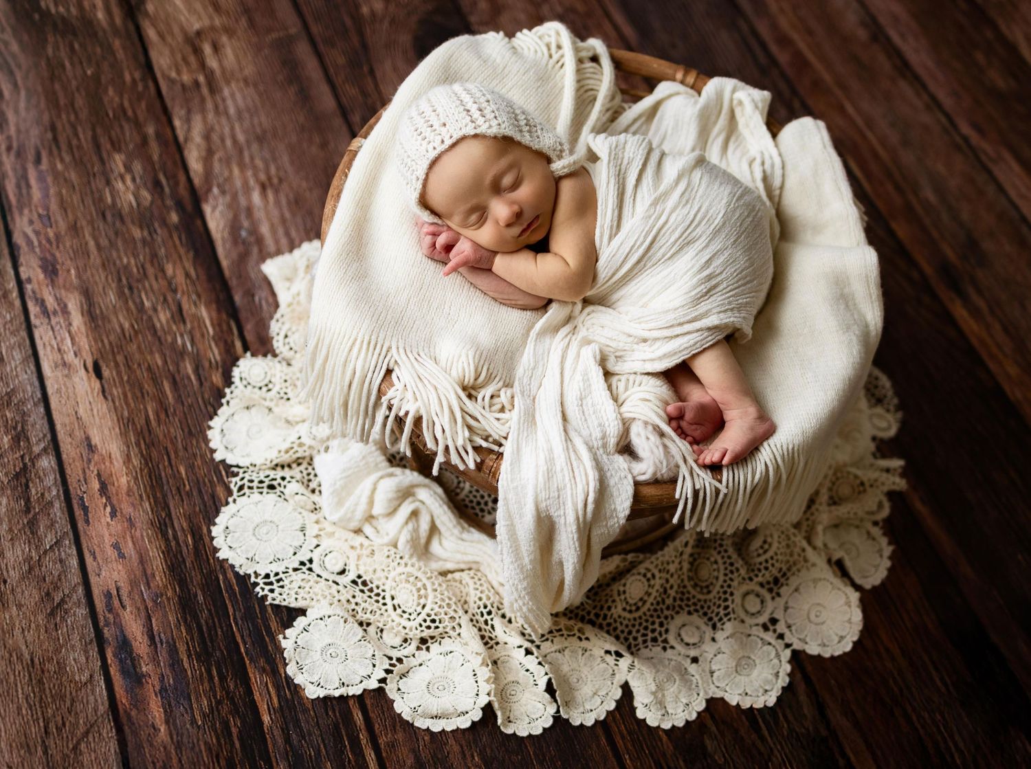 A sleeping newborn baby wrapped in white swaddle cloth lies on vintage lace doilies against dark wooden flooring.