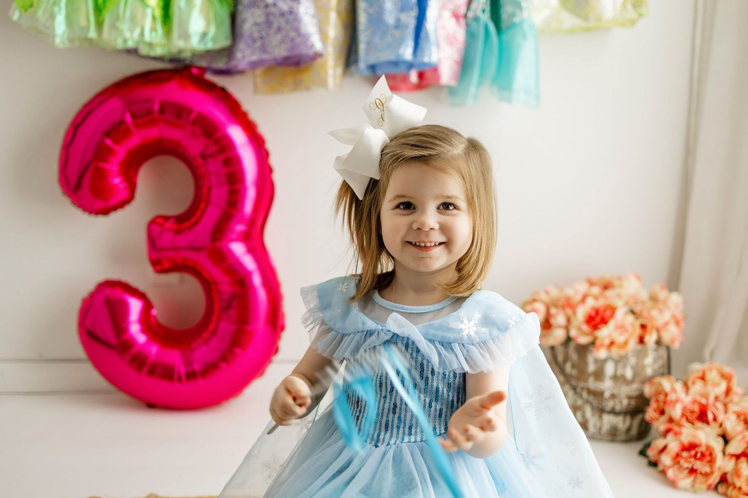 A child stands in front of pink balloon numbers and coral flowers at a birthday celebration.