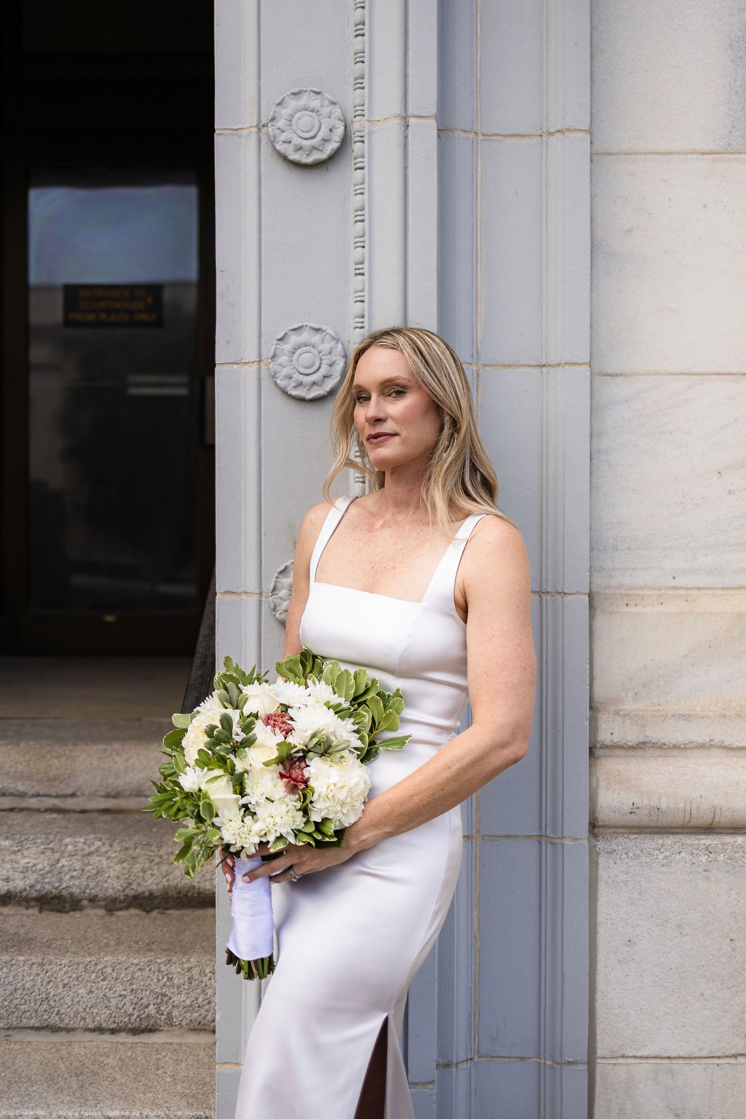 Woman in white dress stands against stone pillar holding elegant white and burgundy wedding bouquet.